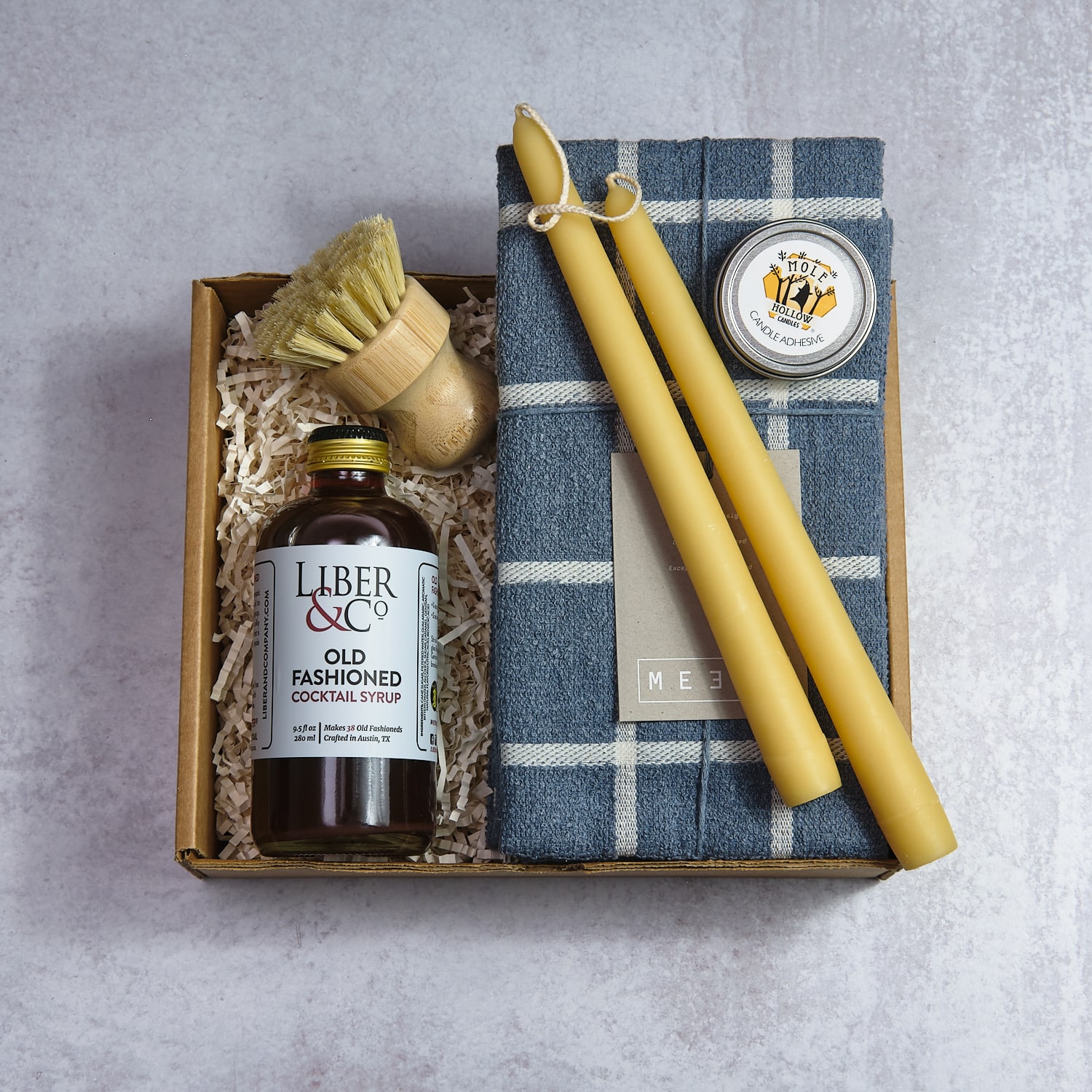 Gift box with yellow candles, a jar of cocktail syrup, and a small container, on a gray surface.