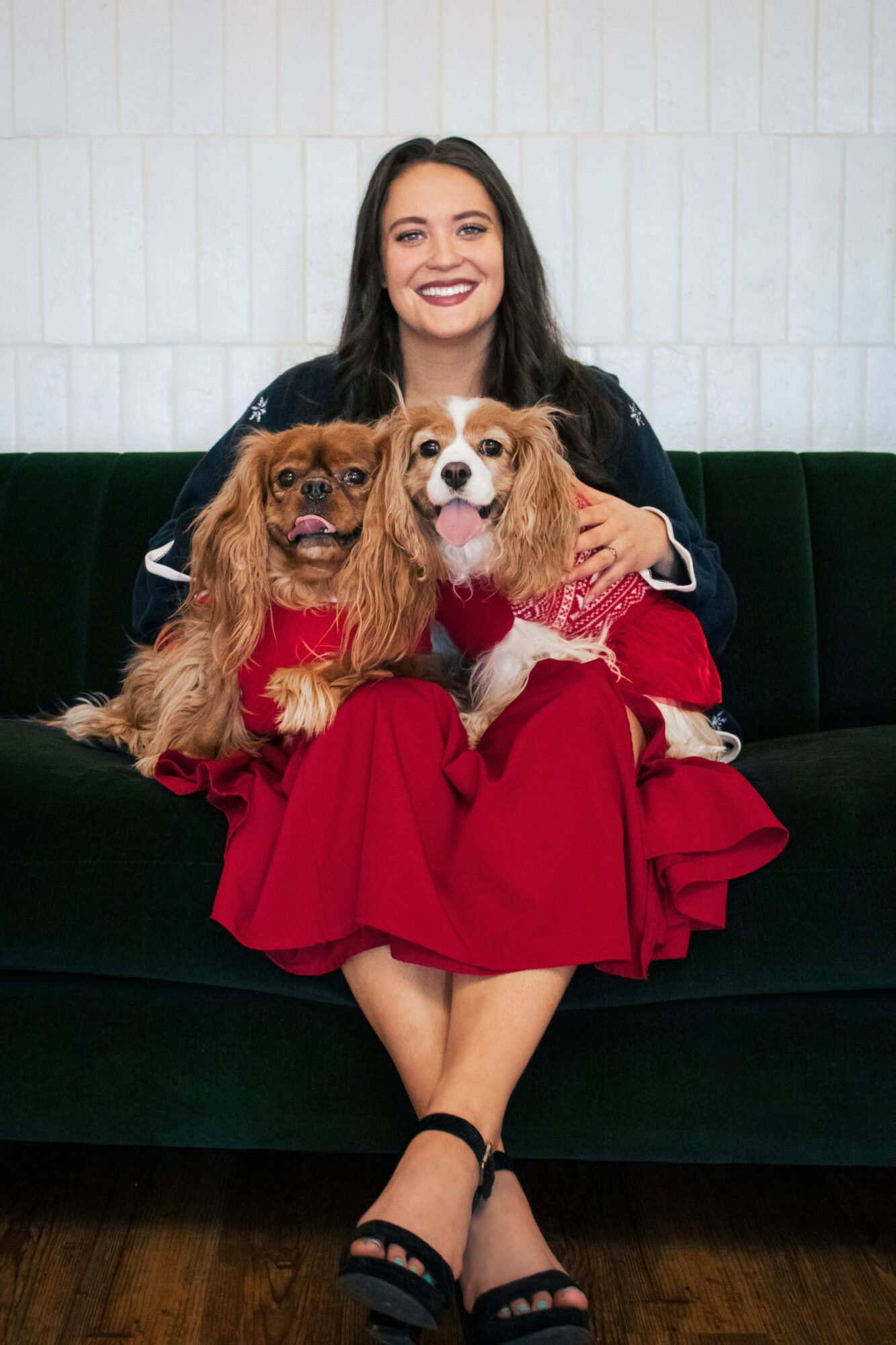 Woman sitting on a dark green sofa holding two small dogs, smiling, with a white wall background.