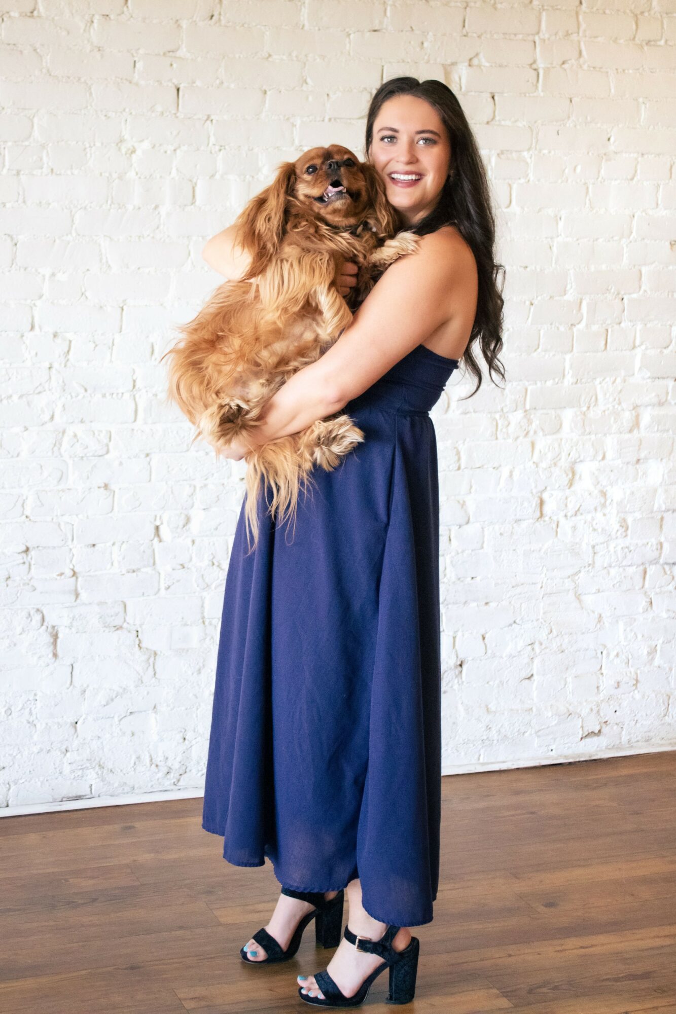 Woman in a blue dress holding a small brown dog against a white brick wall, smiling.