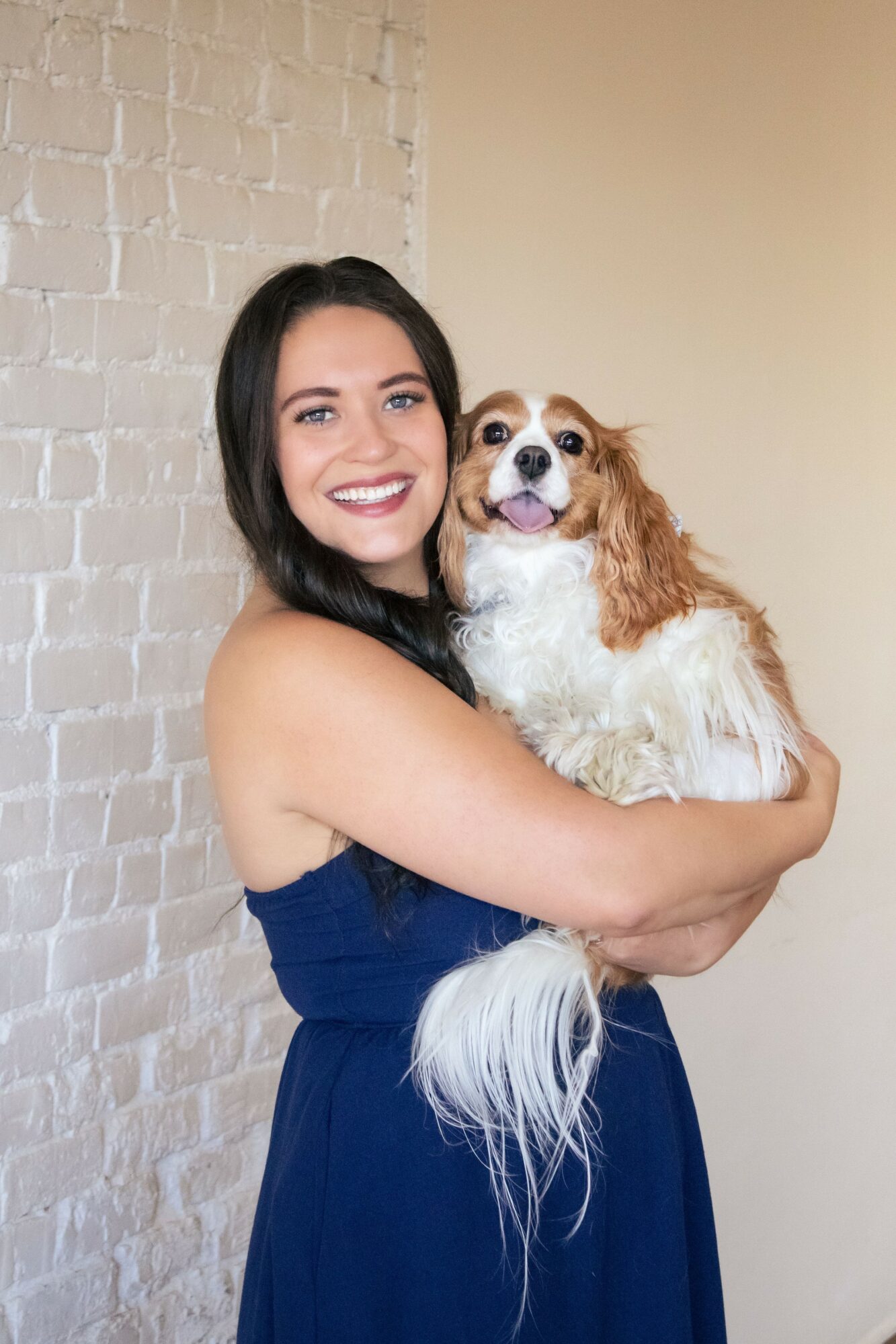 Smiling woman holding a small dog with long ears and white and brown fur, standing against a brick wall.
