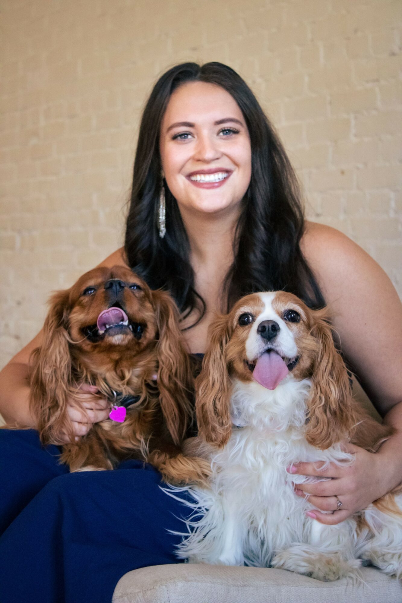 Smiling woman with long dark hair holding two dogs, one with brown fur and the other with white and brown fur, against a beige brick wall.