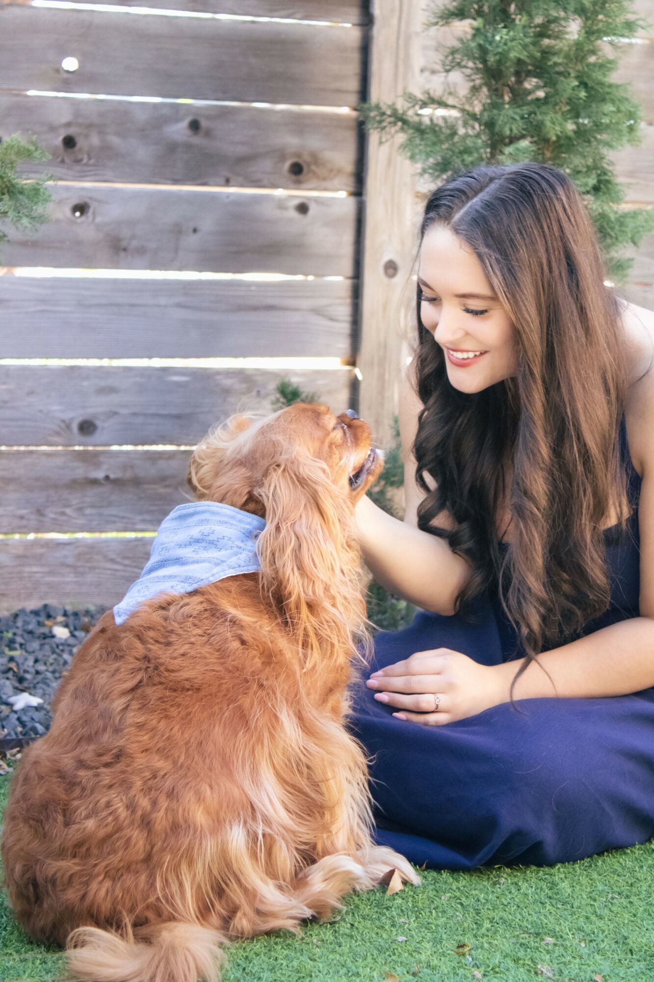Young woman with long dark hair smiling at a sitting golden retriever outdoors, wooden fence and greenery in background.