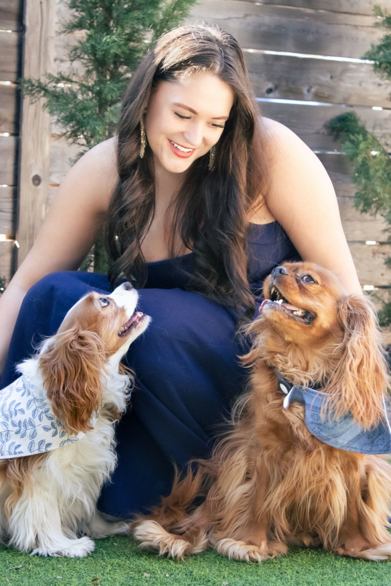 Young woman with long dark hair smiling, sitting outdoors with two dogs, one small and one large, on grass.