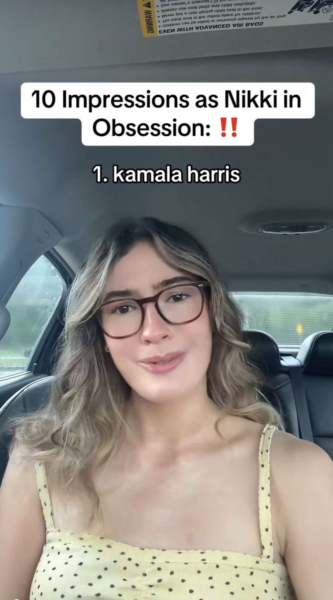 Young woman with glasses and wavy hair sitting in a car, wearing a yellow polka dot top, smiling.