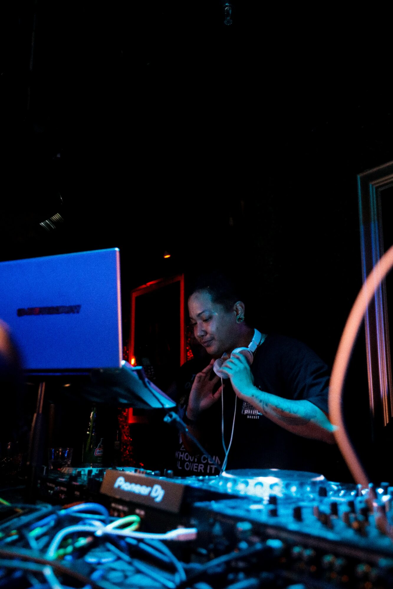 DJ wearing headphones operates equipment in a dark club with blue and red lighting, surrounded by electronic devices and cables.