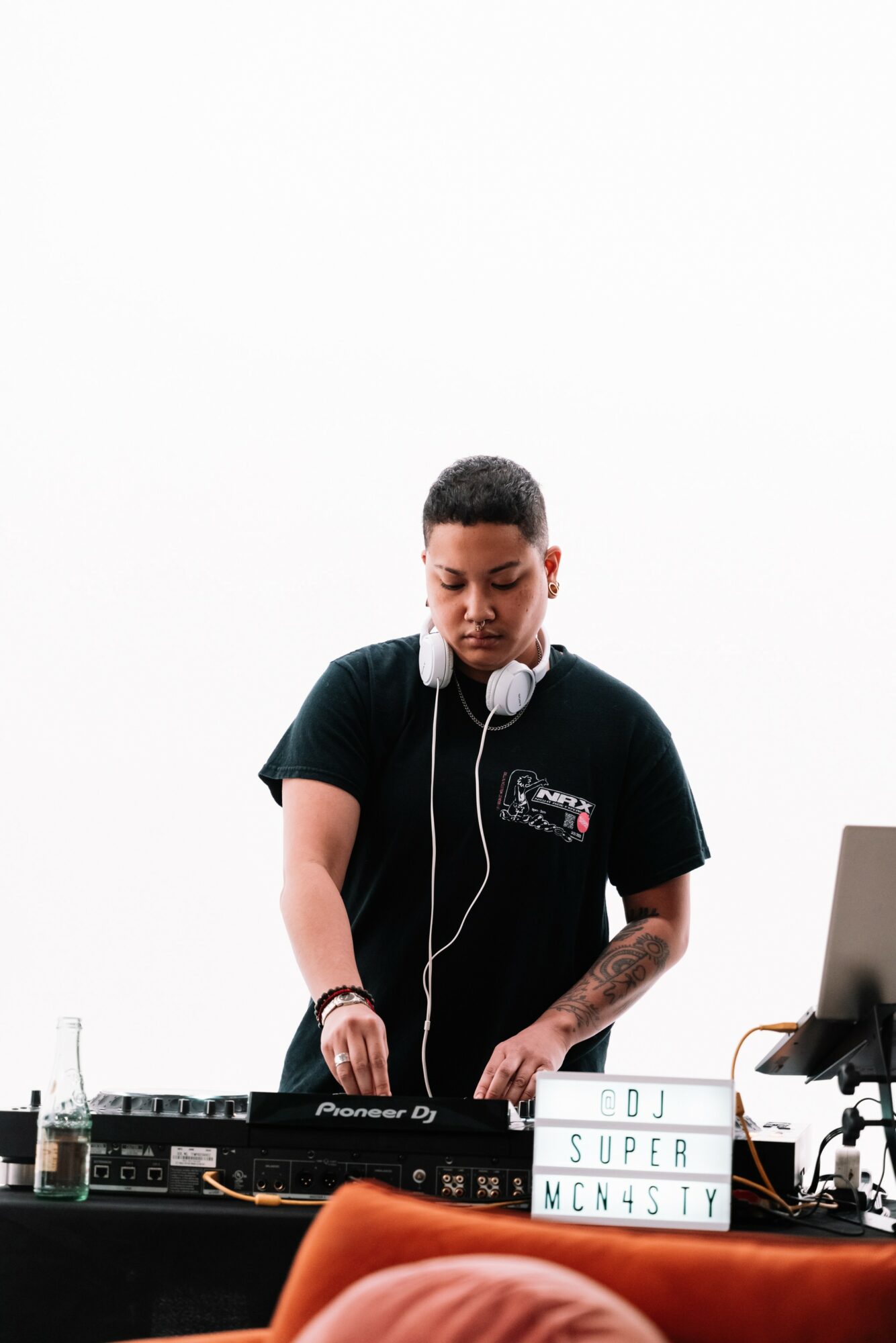 Young man with headphones operating DJ equipment, with a laptop and a lightbox displaying text, against a white background.