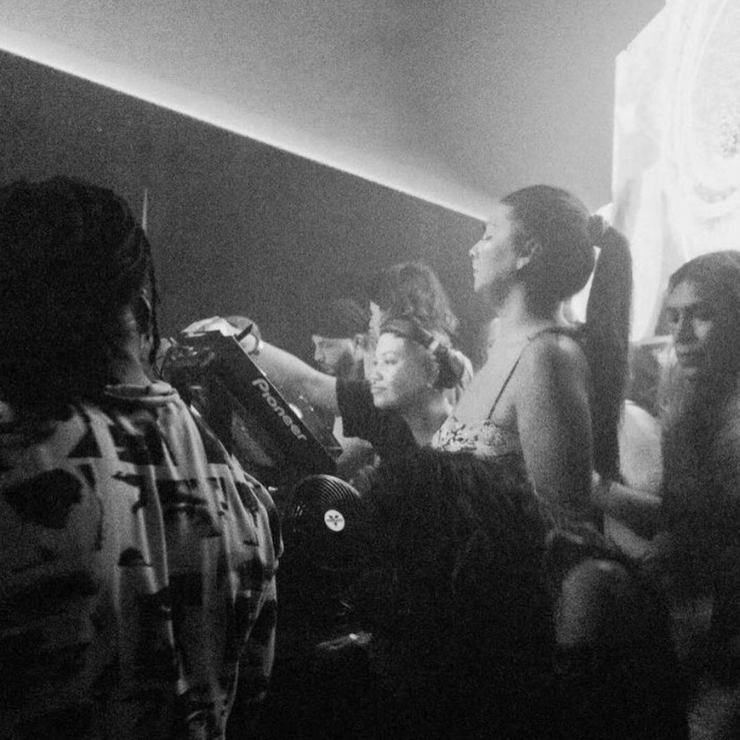 Group of people at a DJ booth, some with headphones, in a dimly lit setting.