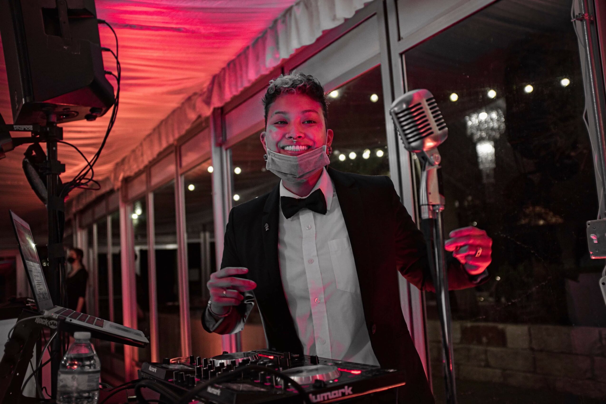 Young person in formal attire with a mask, standing near a microphone at an event, with a window and city lights behind.
