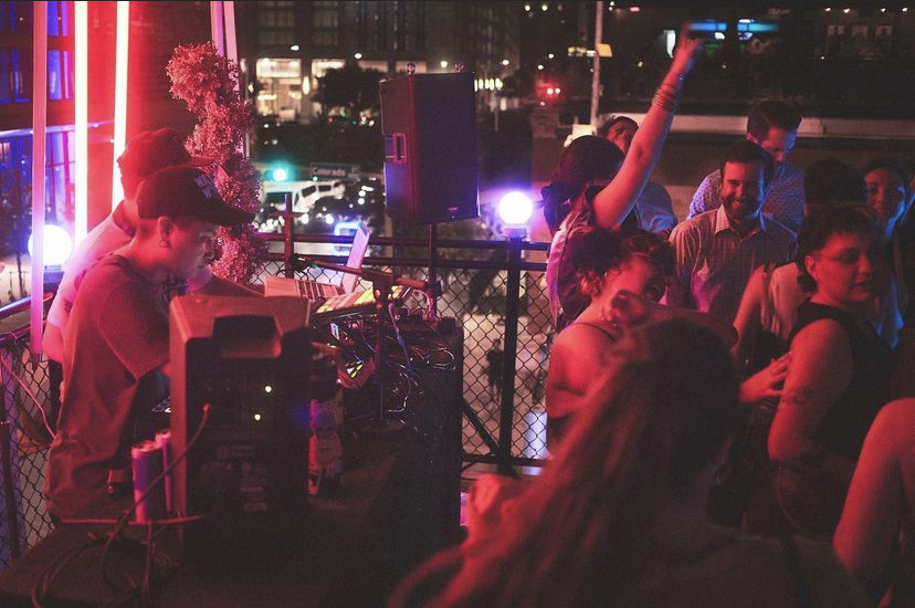 People dancing and socializing at a nighttime outdoor party with colorful lights and city background.