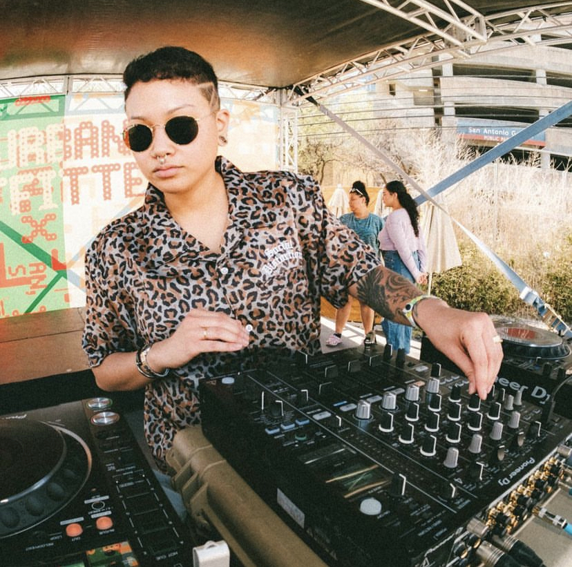 Person wearing leopard print shirt and sunglasses operating DJ equipment outdoors with people in background.
