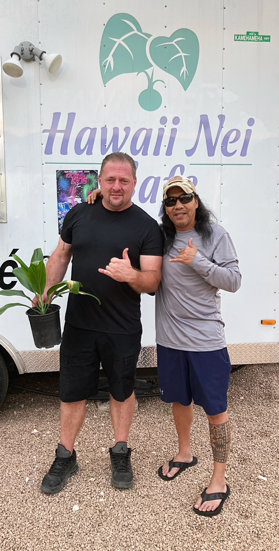 Two people standing in front of a white wall with Hawaii Nei sign, smiling, one making a shaka gesture, outdoors.