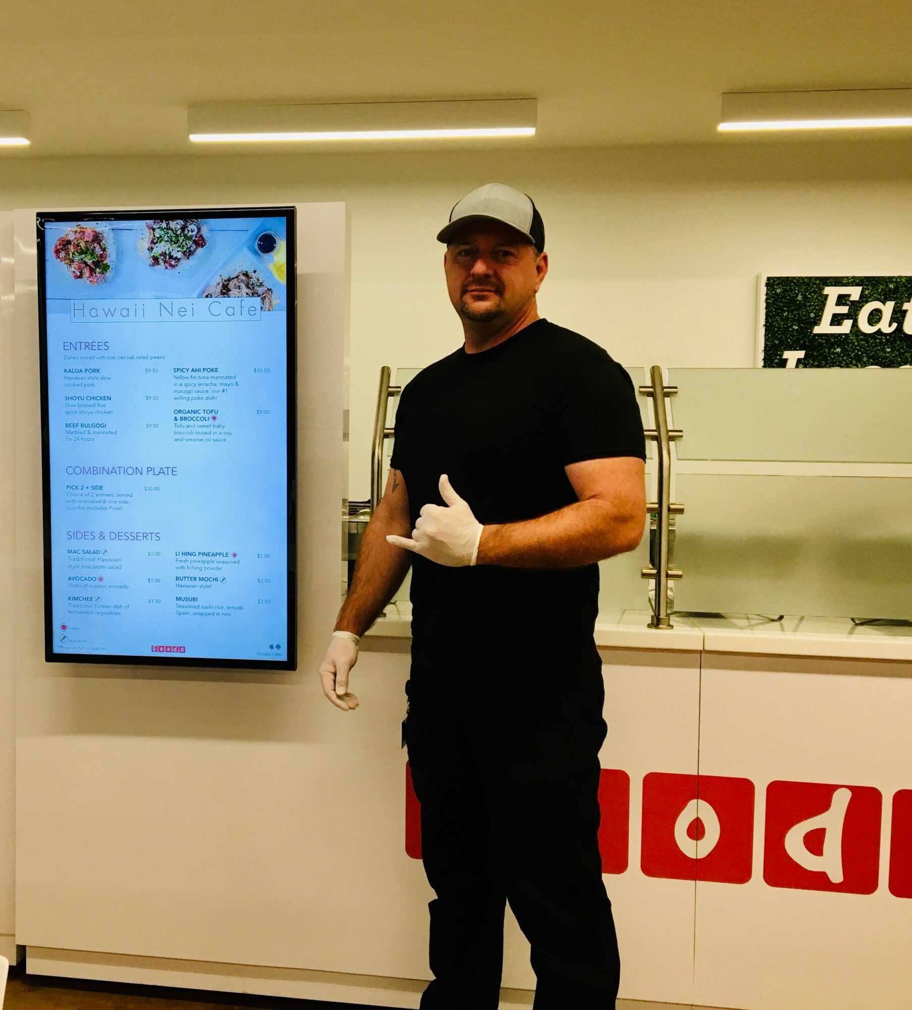 Man wearing black shirt, white gloves, and a cap standing indoors near a digital display and a counter.