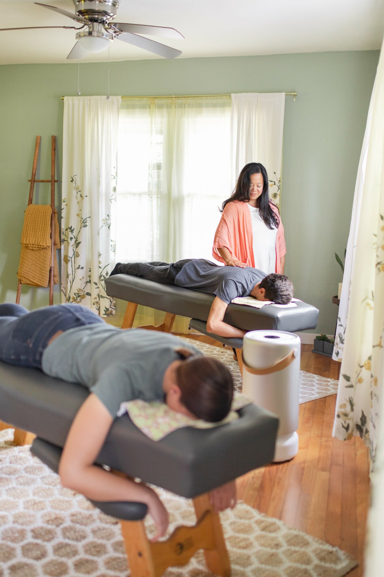 Person lying on a massage table with a towel, another person standing nearby in a room with light green walls and large windows.