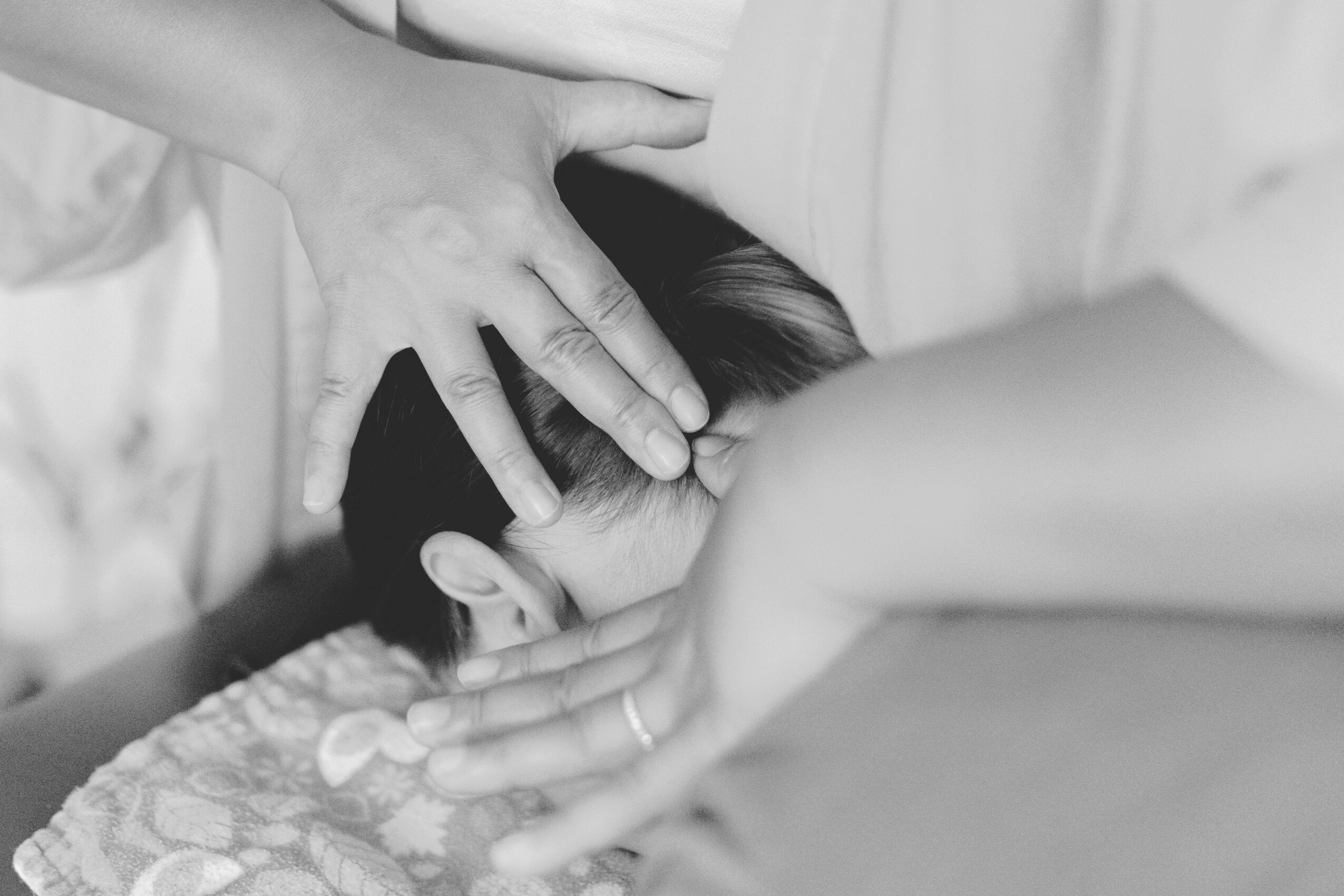 Two people hold a child's head gently, with one hand on the forehead and the other on the back of the head, in black and white.