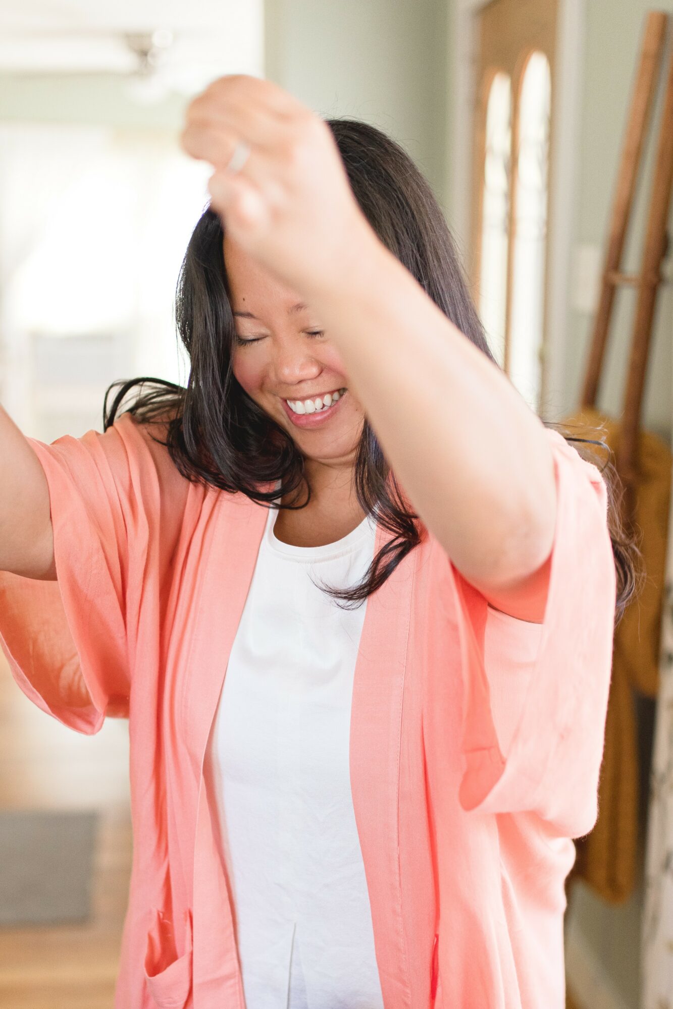 Woman smiling and laughing, wearing a white shirt and coral-colored cardigan, indoors with sunlight and wooden furniture.