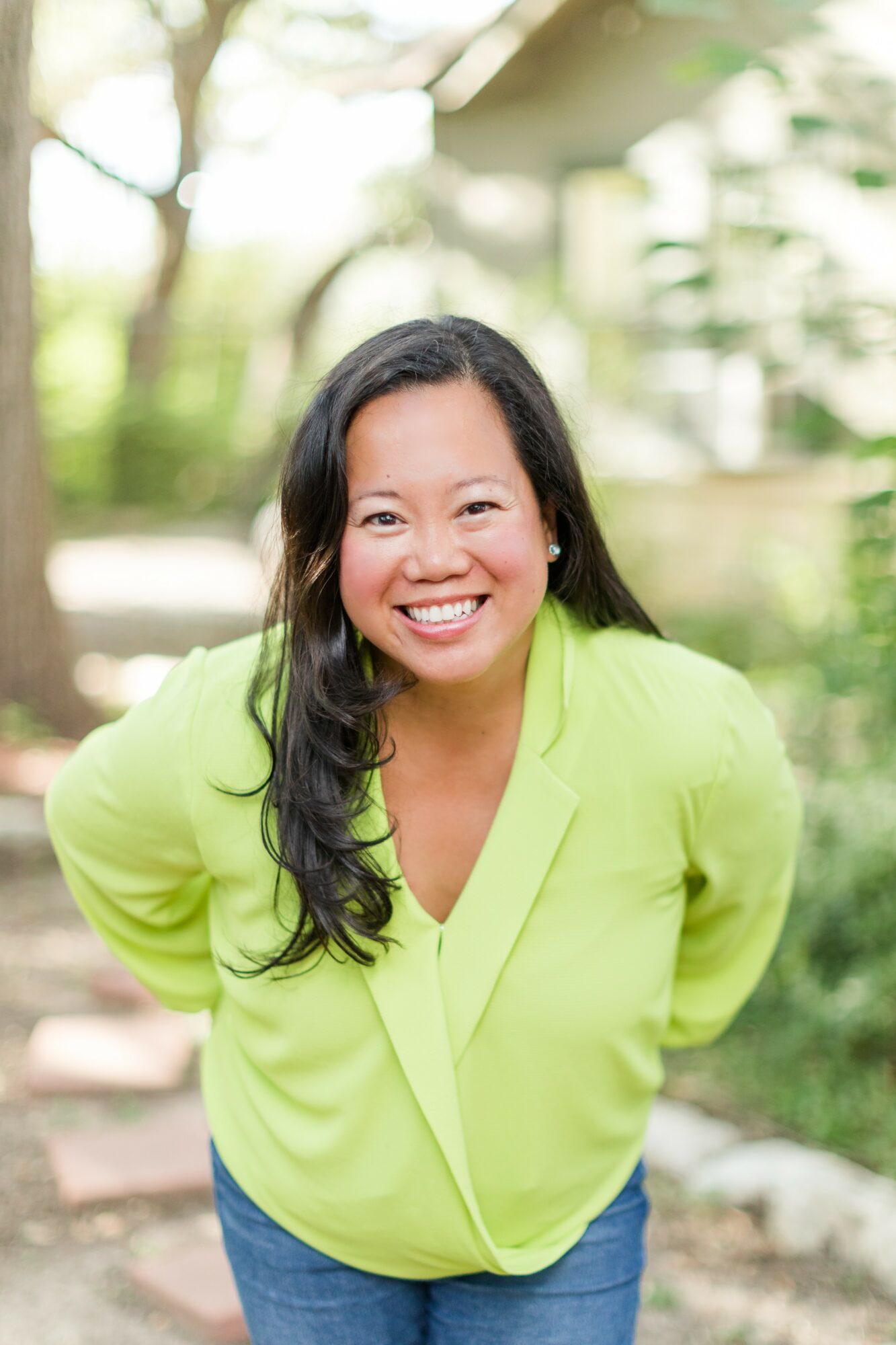 Smiling woman with long dark hair in a bright green blouse outdoors, greenery in background.
