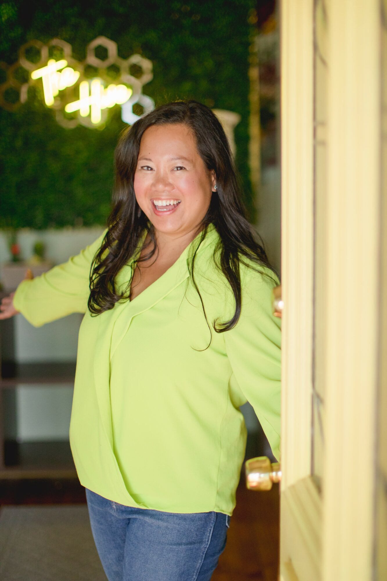 Woman with long dark hair smiling, wearing a bright yellow top, standing indoors near a door.