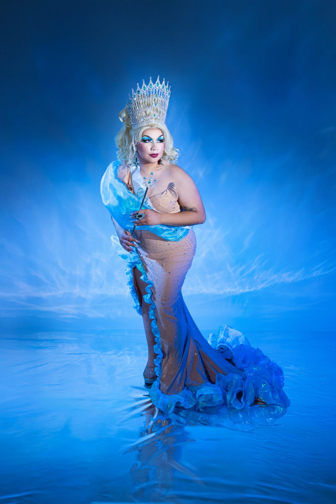Person in elaborate costume with crown, standing in water, surrounded by blue lighting and mist, looking at camera.