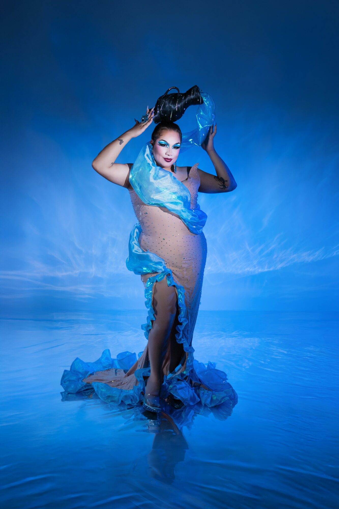 A woman in a flowing blue dress poses with her arms raised, standing on a reflective surface with a blue background.