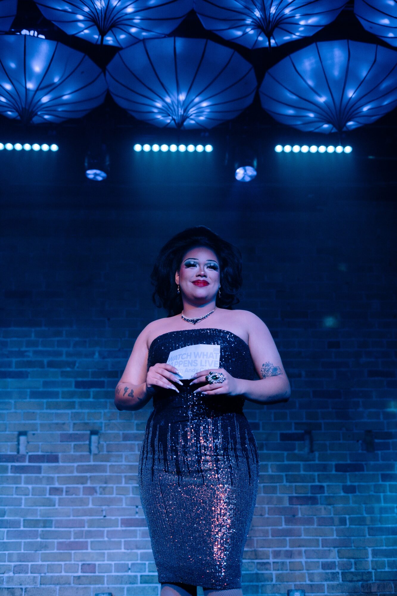 Woman in a sparkly dress holding a card, standing on stage with blue lighting and umbrella-shaped ceiling decorations.