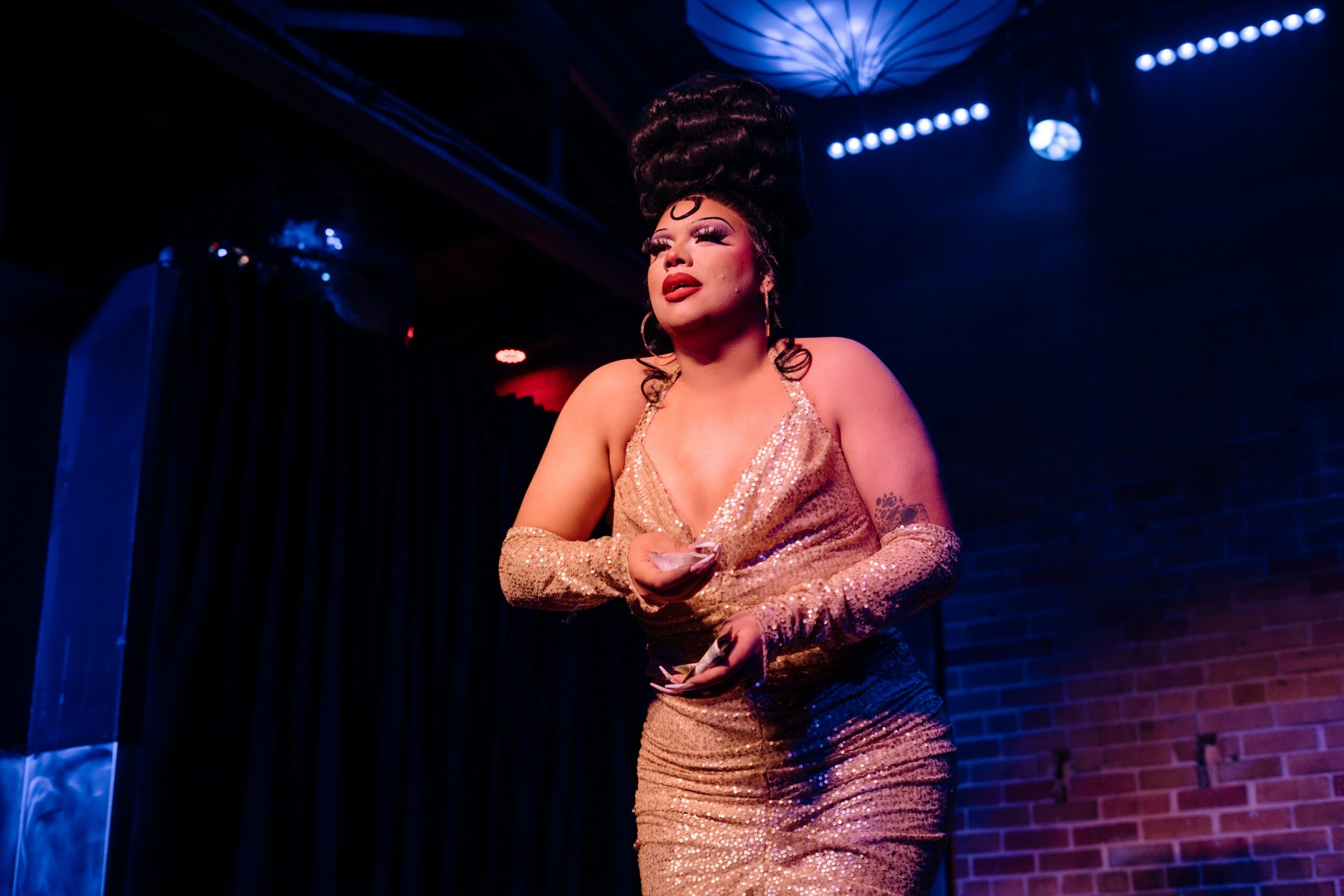 Performer with styled hair and glamorous outfit on stage, holding a microphone, with stage lights and brick wall background.