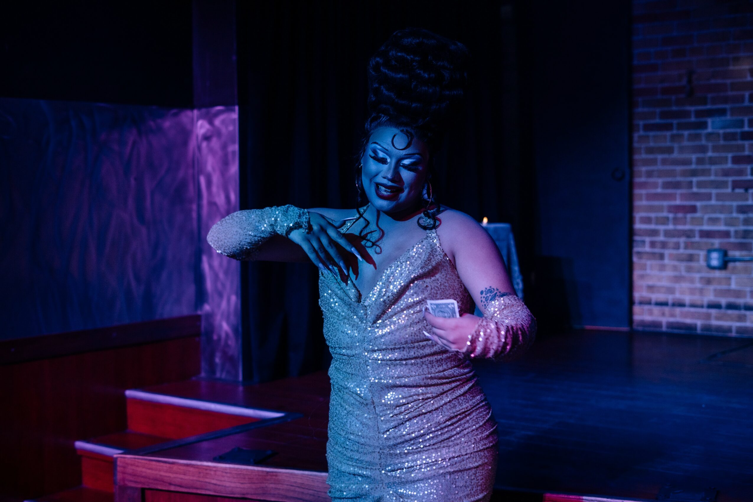 Performer in a shiny dress with elaborate hairstyle on stage, holding a phone, with a brick wall background.