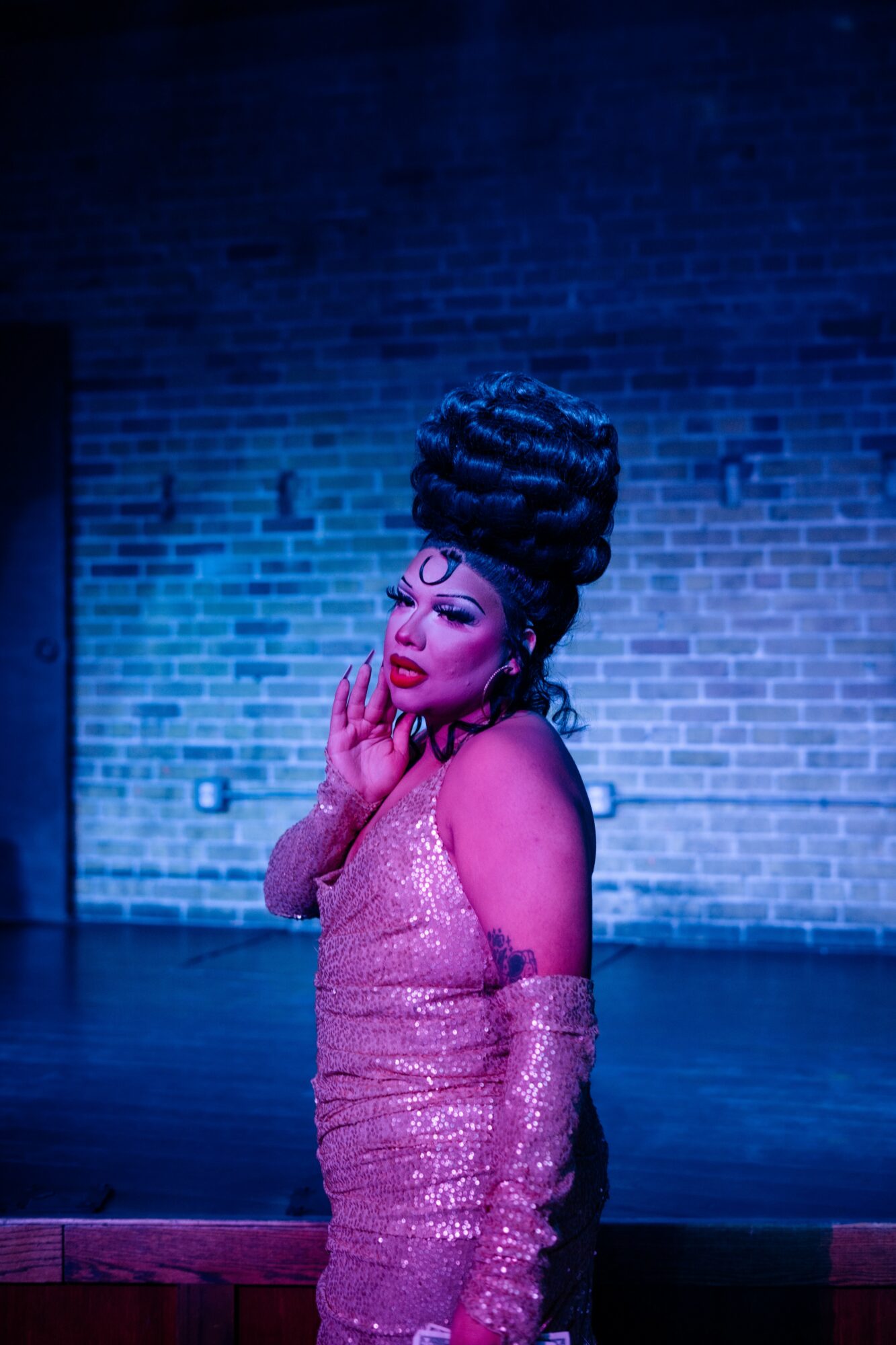 Woman with elaborate braided hairstyle and pink dress stands against a brick wall, eyes closed, hands near face.