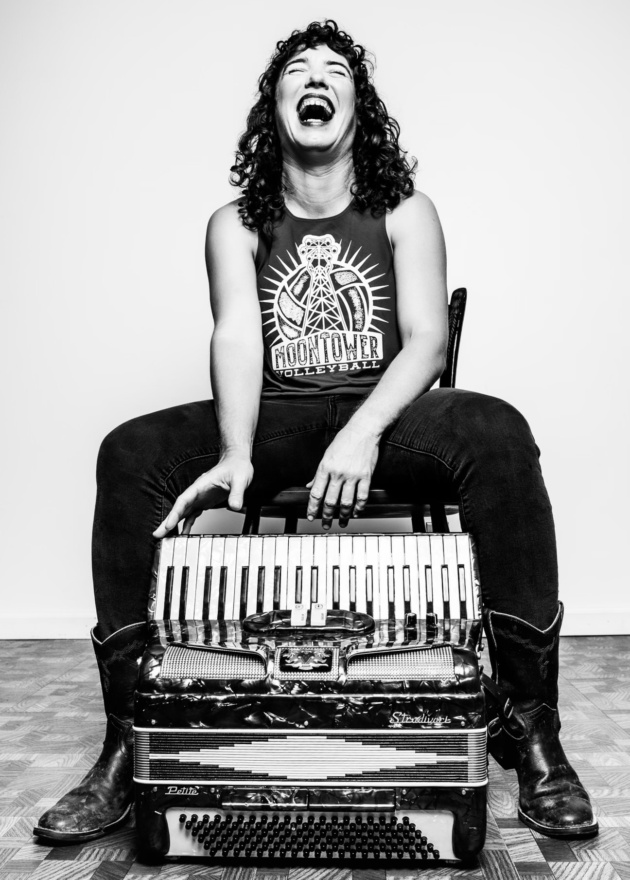 Person with curly hair laughing, sitting on a suitcase with a keyboard, wearing a sleeveless shirt and boots.