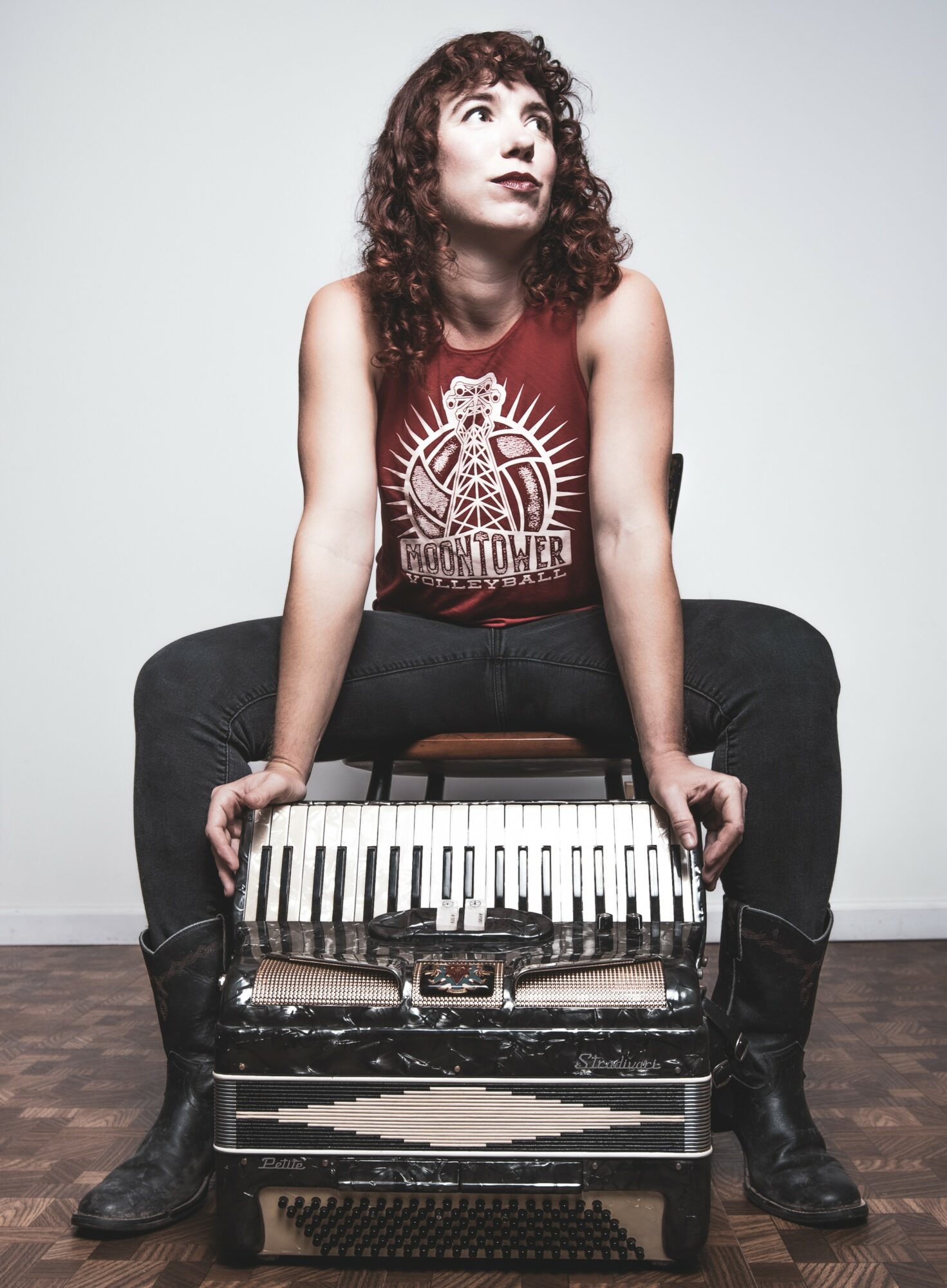 Woman with curly hair in a red sleeveless shirt sitting on a piano, legs apart, hands on piano, against a plain white wall.