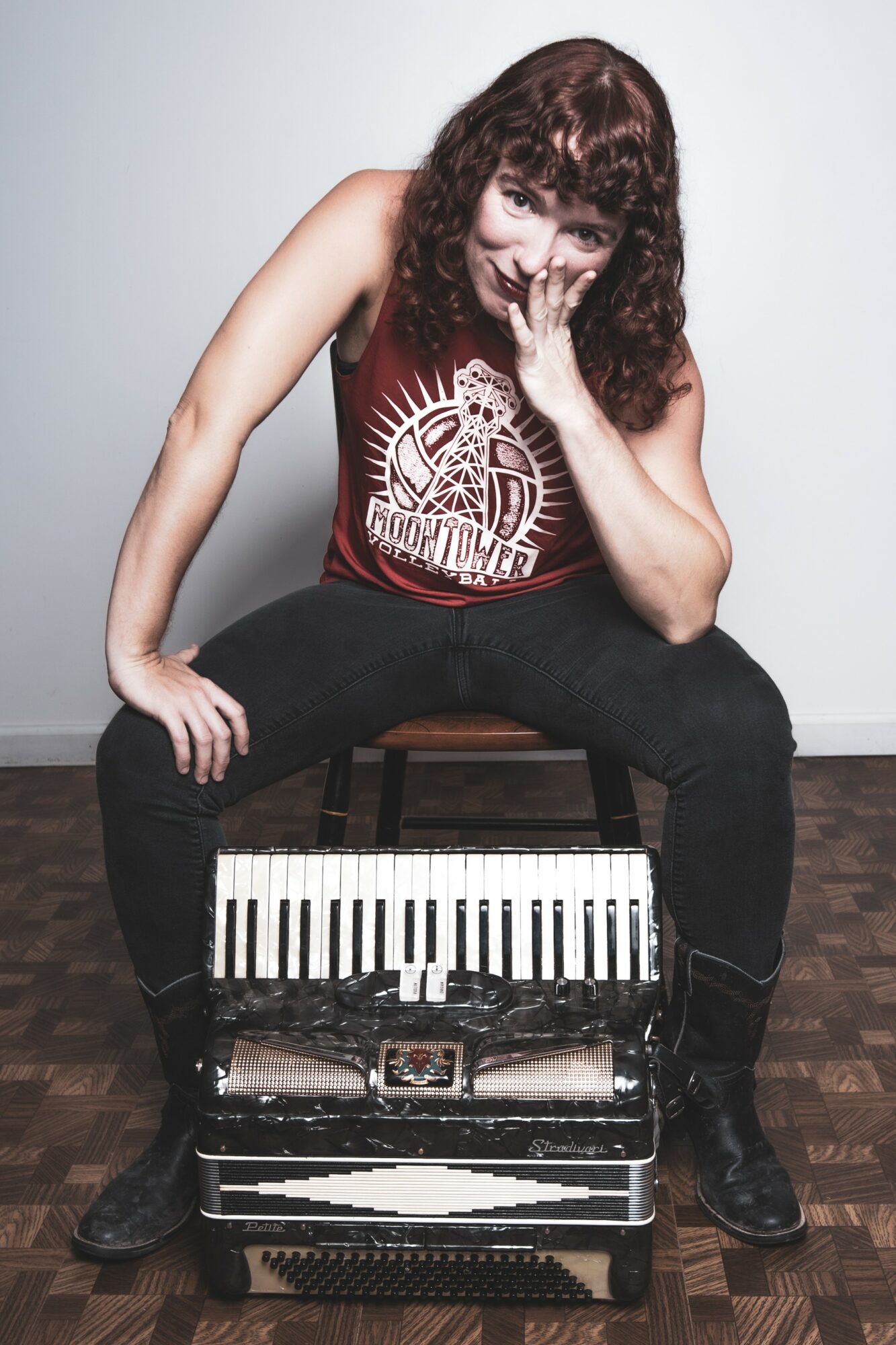Person with curly hair sitting on a stool, covering mouth with hand, wearing a red tank top and black pants, with a keyboard in front.