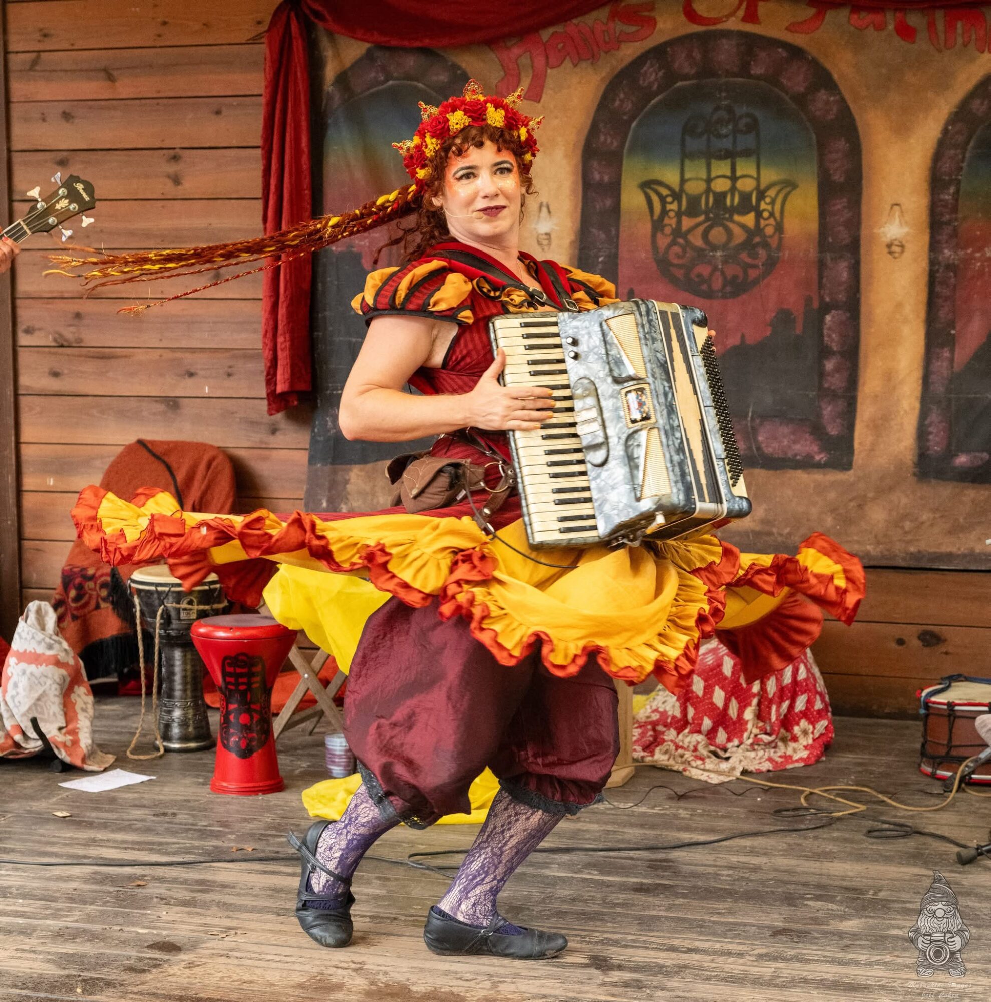Woman in colorful costume playing an accordion on stage with wooden background and props.