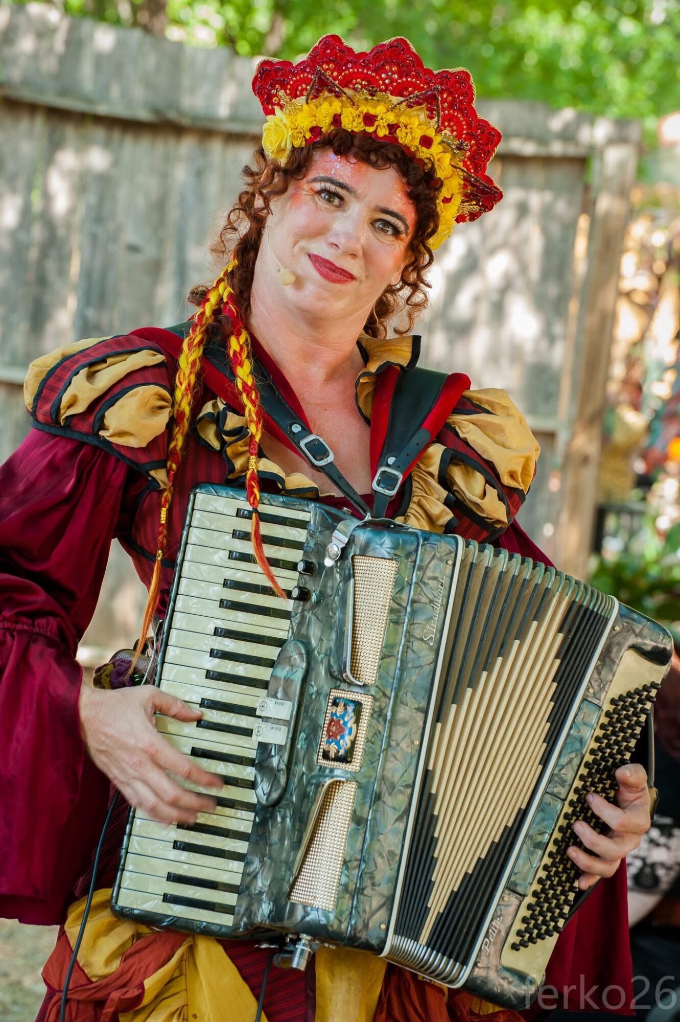 Woman in colorful traditional costume playing an accordion outdoors, wearing a floral headpiece, smiling.