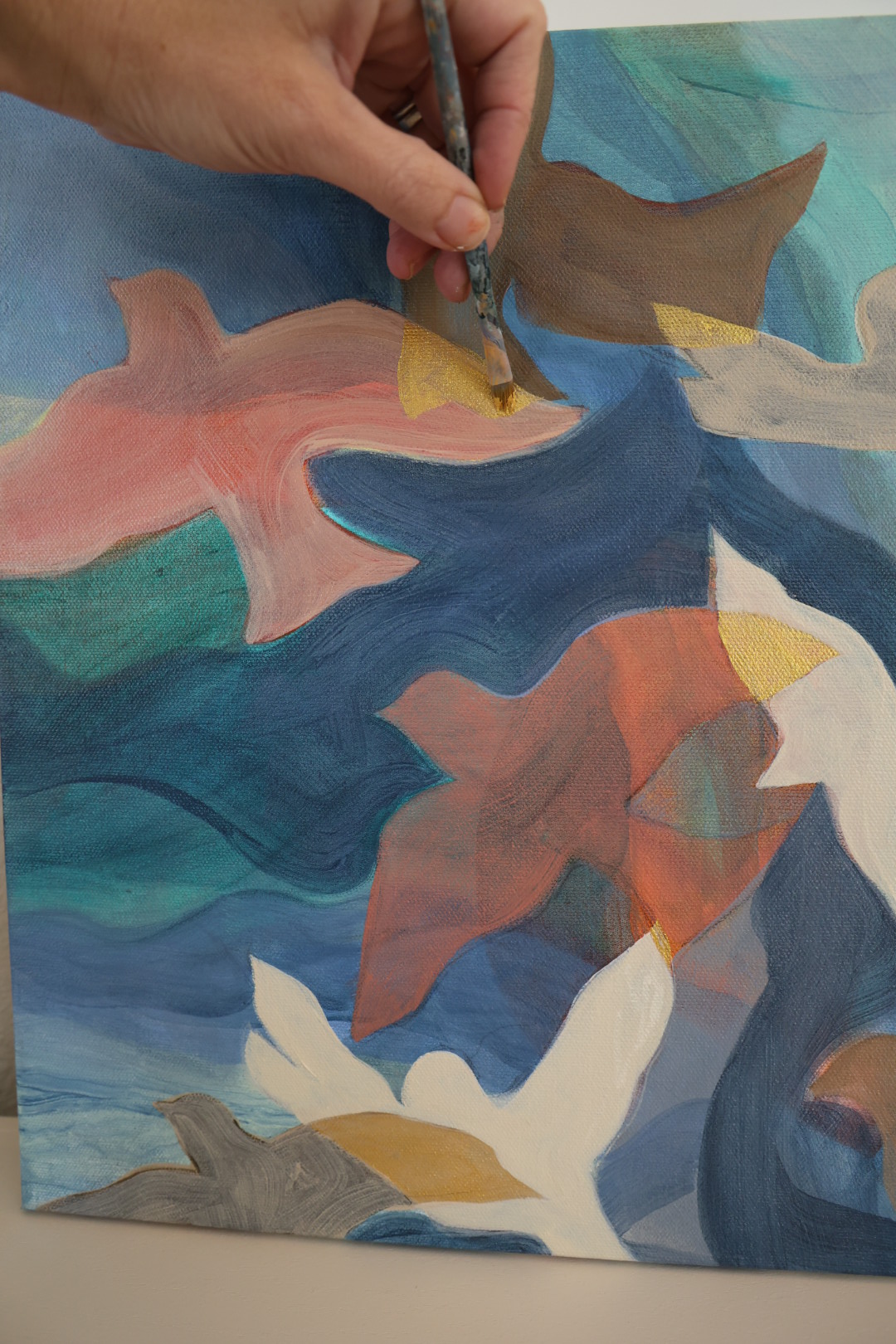 Hand holding a paintbrush painting abstract shapes with blue, brown, yellow, and white colors on a canvas.