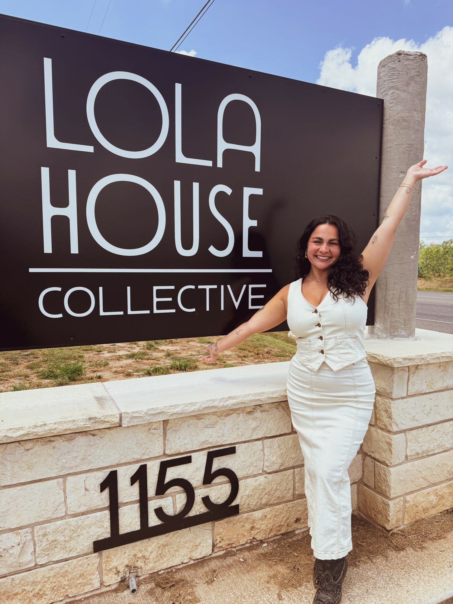 Woman with curly hair in a white dress smiling and gesturing with one arm in front of a sign reading 'LOLA HOUSE COLLECTIVE' and the number 1155.