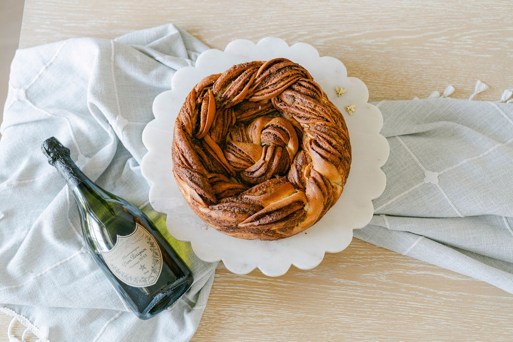 A round, braided pastry on a white scalloped plate, with a bottle of sparkling wine and a light-colored cloth nearby.