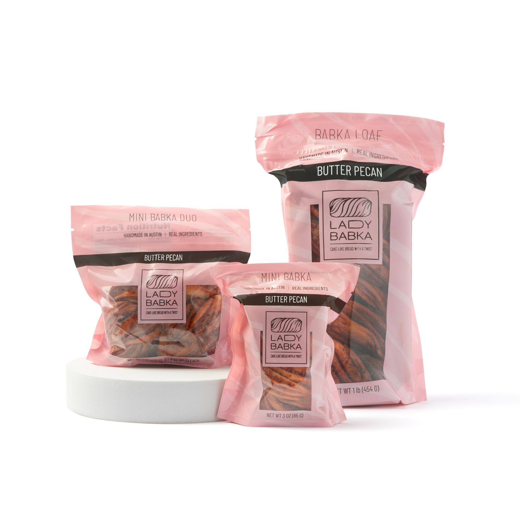 Three pink and black packages of Lady Babka Butter Pecan flavor, with one open package showing baked goods, on a white surface.