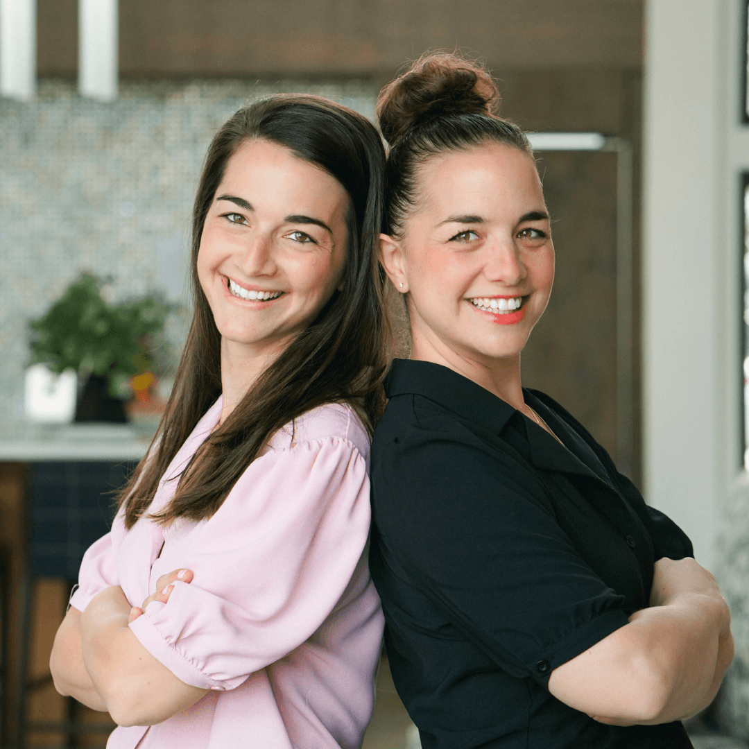 Two women standing close together, smiling, with arms crossed, in an indoor setting with plants and a window behind them.