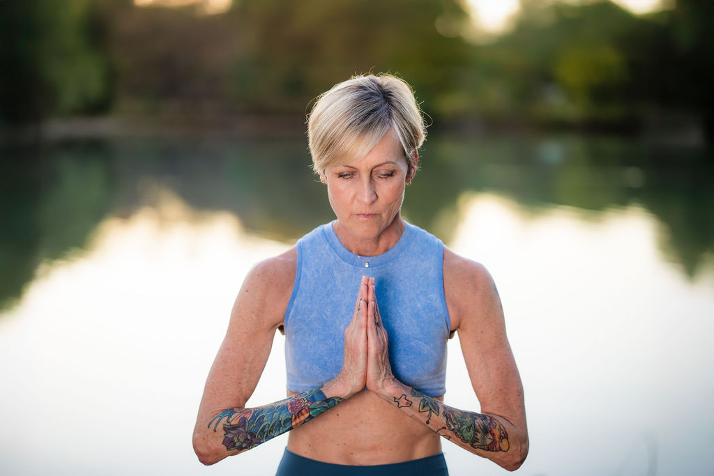 Woman with short gray hair practicing yoga outdoors, hands in prayer position, wearing a blue sleeveless top, near water.