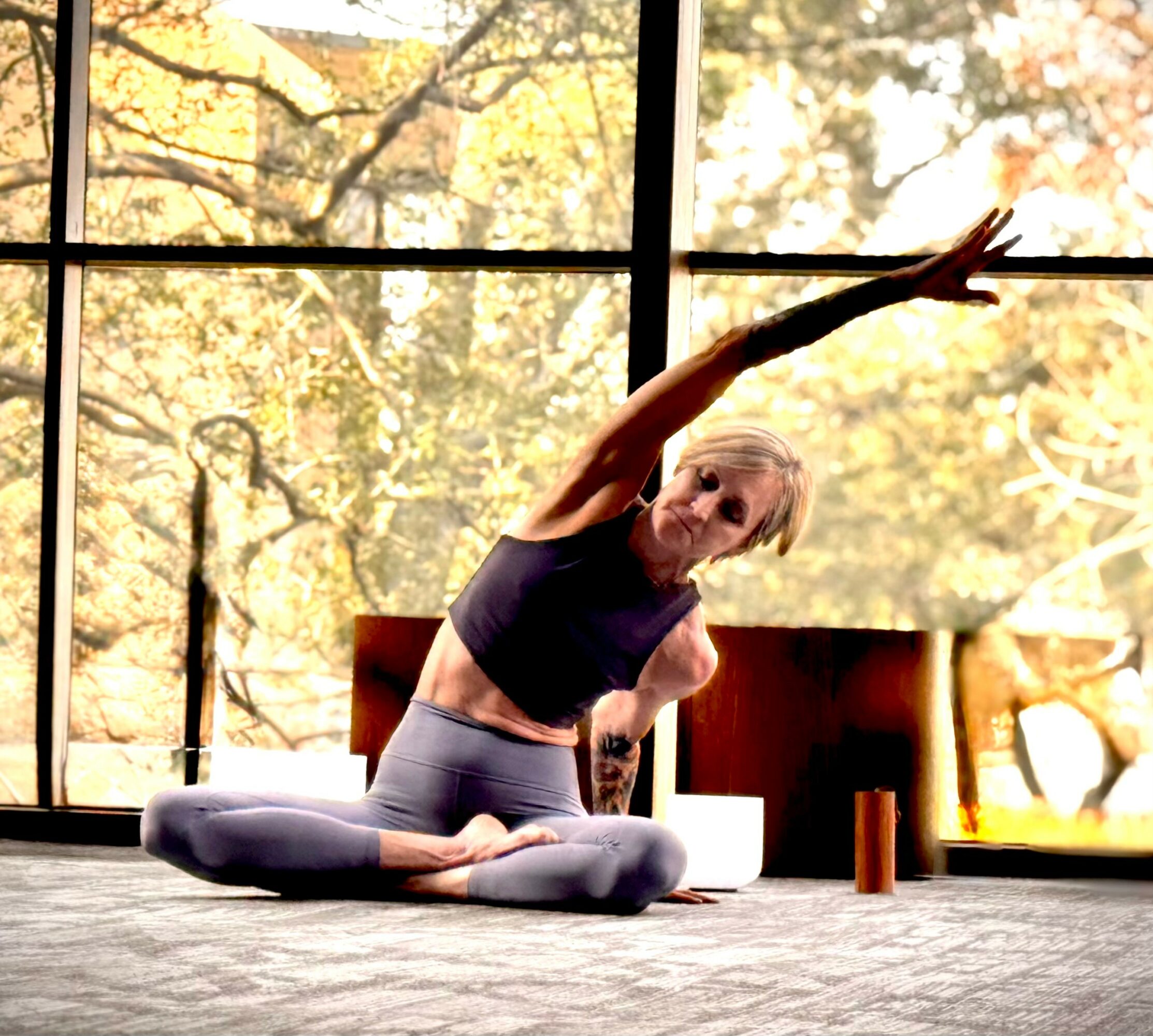 Person practicing yoga indoors with large window showing trees outside, stretching arm upward, sitting cross-legged on mat.