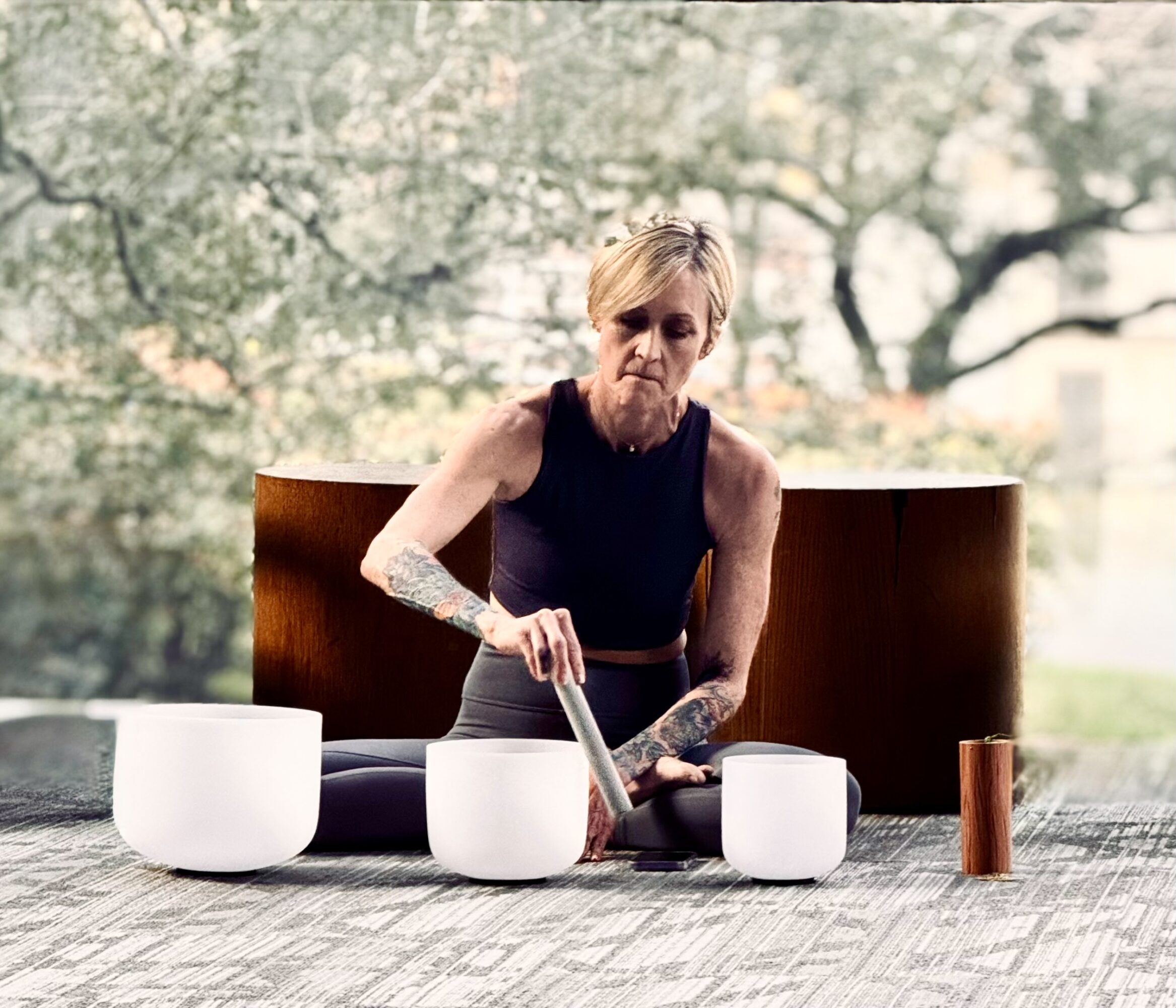 Woman playing singing bowls outdoors, wearing a black sleeveless top, with trees in the background.