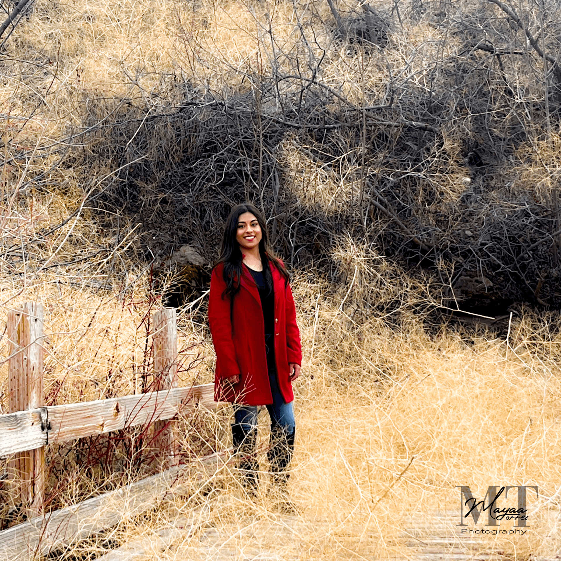 Young woman with long dark hair wearing a red coat standing in a dry grassy field with bushes and trees behind her.