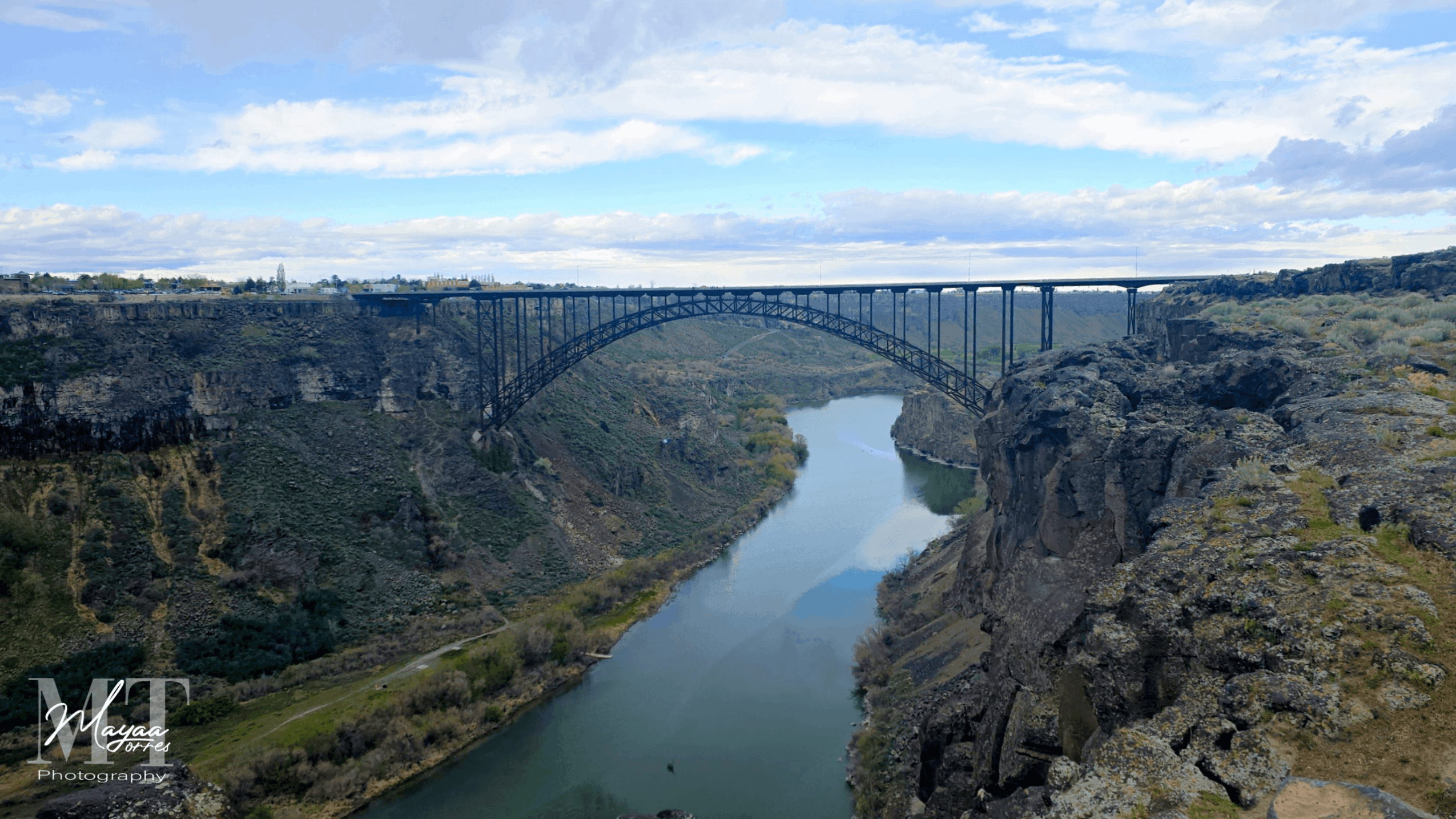 Bridge spanning a river between cliffs with a cloudy sky above.