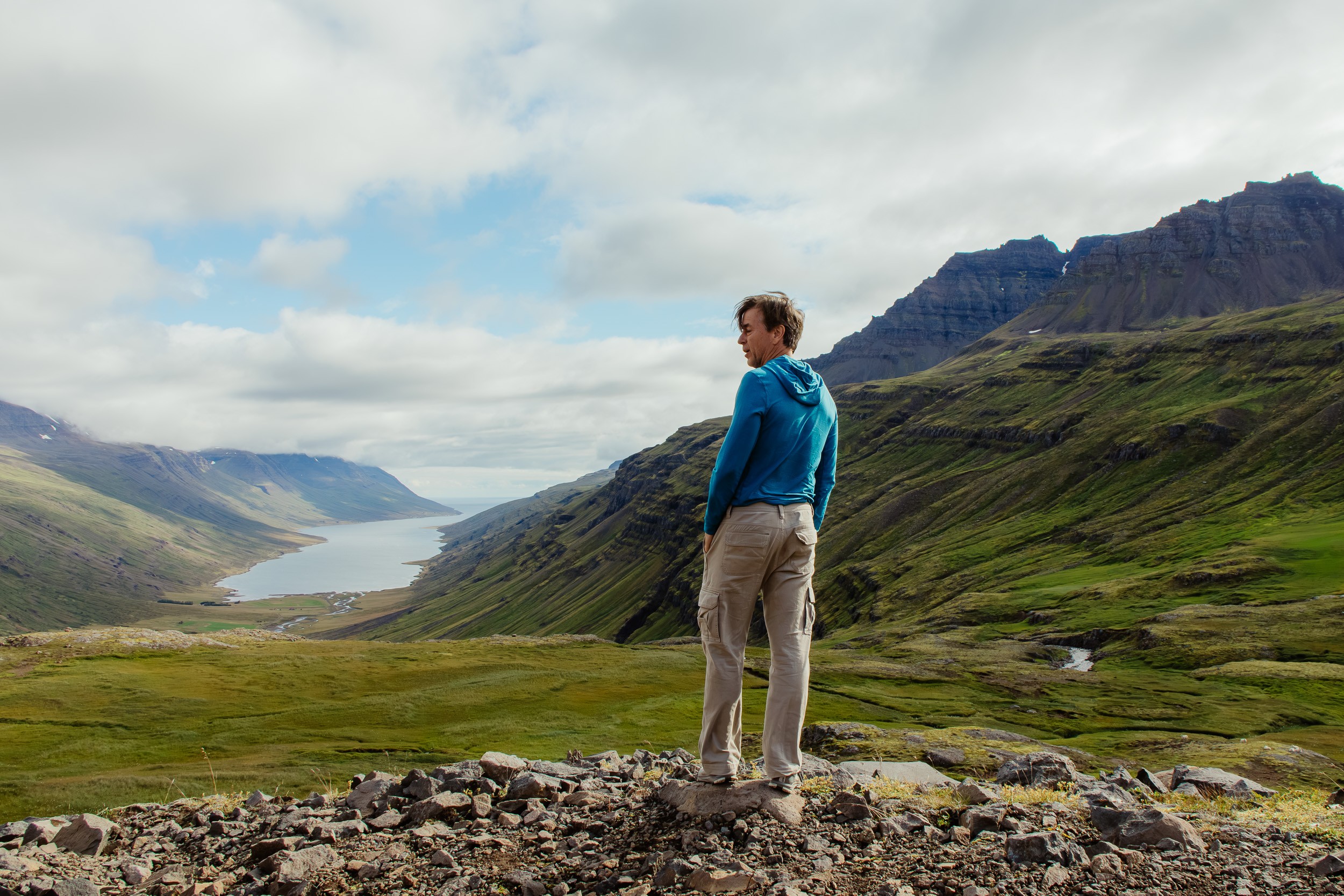 Person standing on rocky ground overlooking green valley and mountains under cloudy sky.