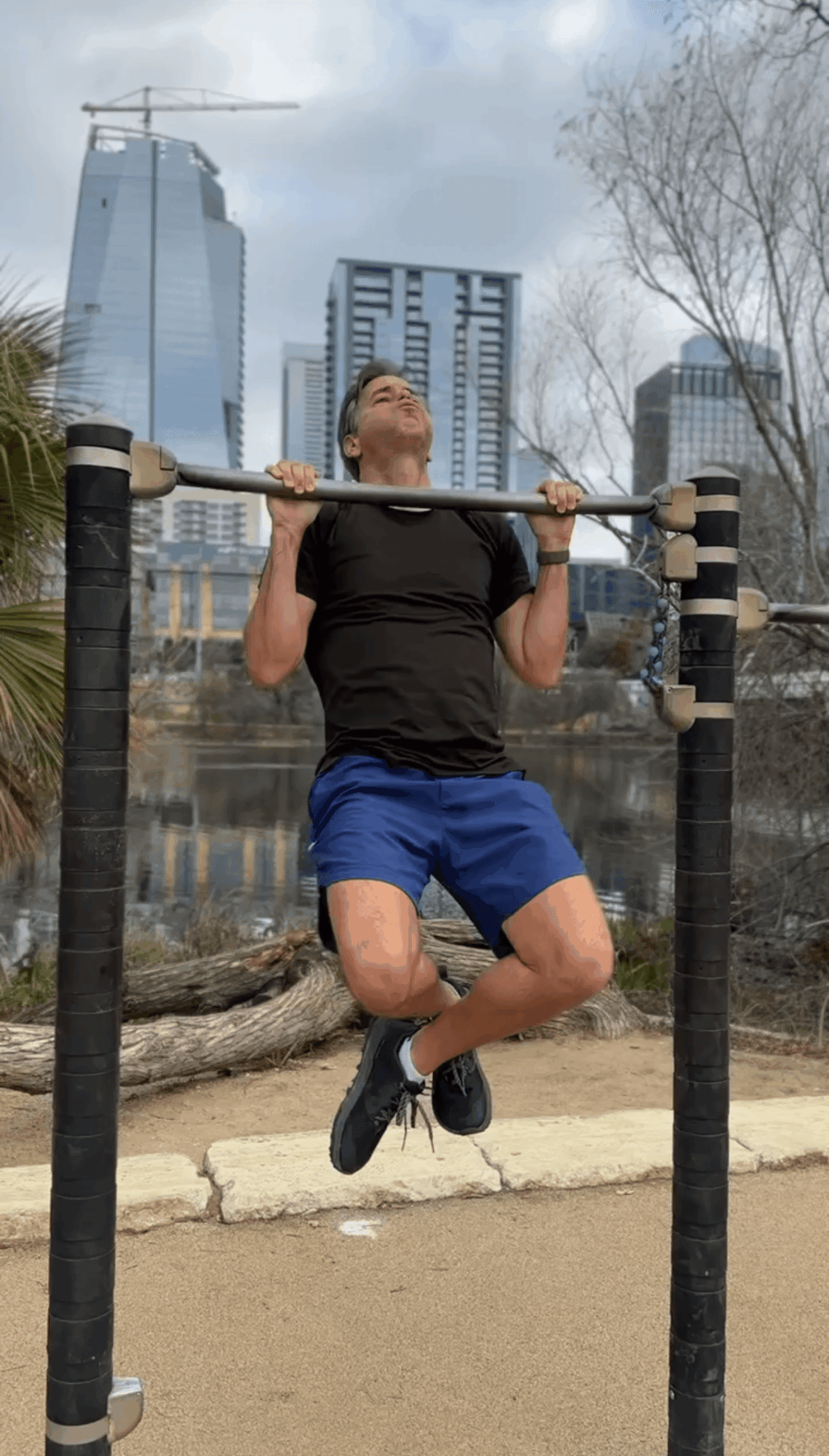 Person performing pull-up exercise outdoors with city buildings in background.