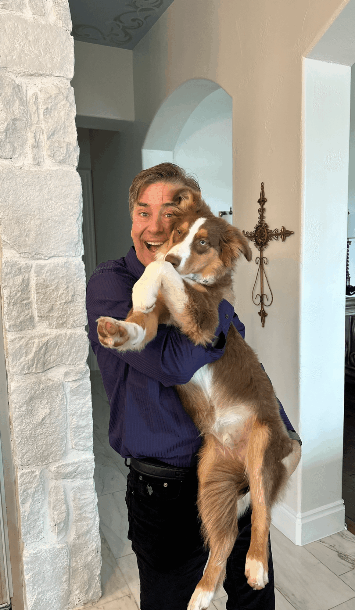 Man holding a large dog indoors, smiling happily, with a decorative wall sconce behind him.