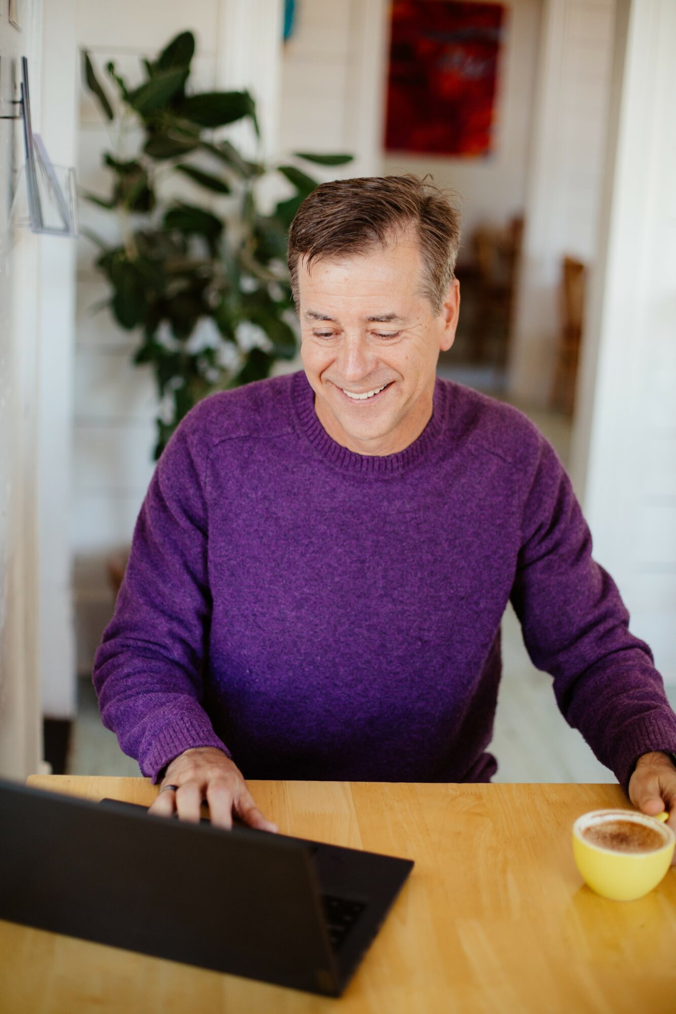 Man in purple sweater smiling while using a laptop at a wooden table, with a plant and a room in the background.
