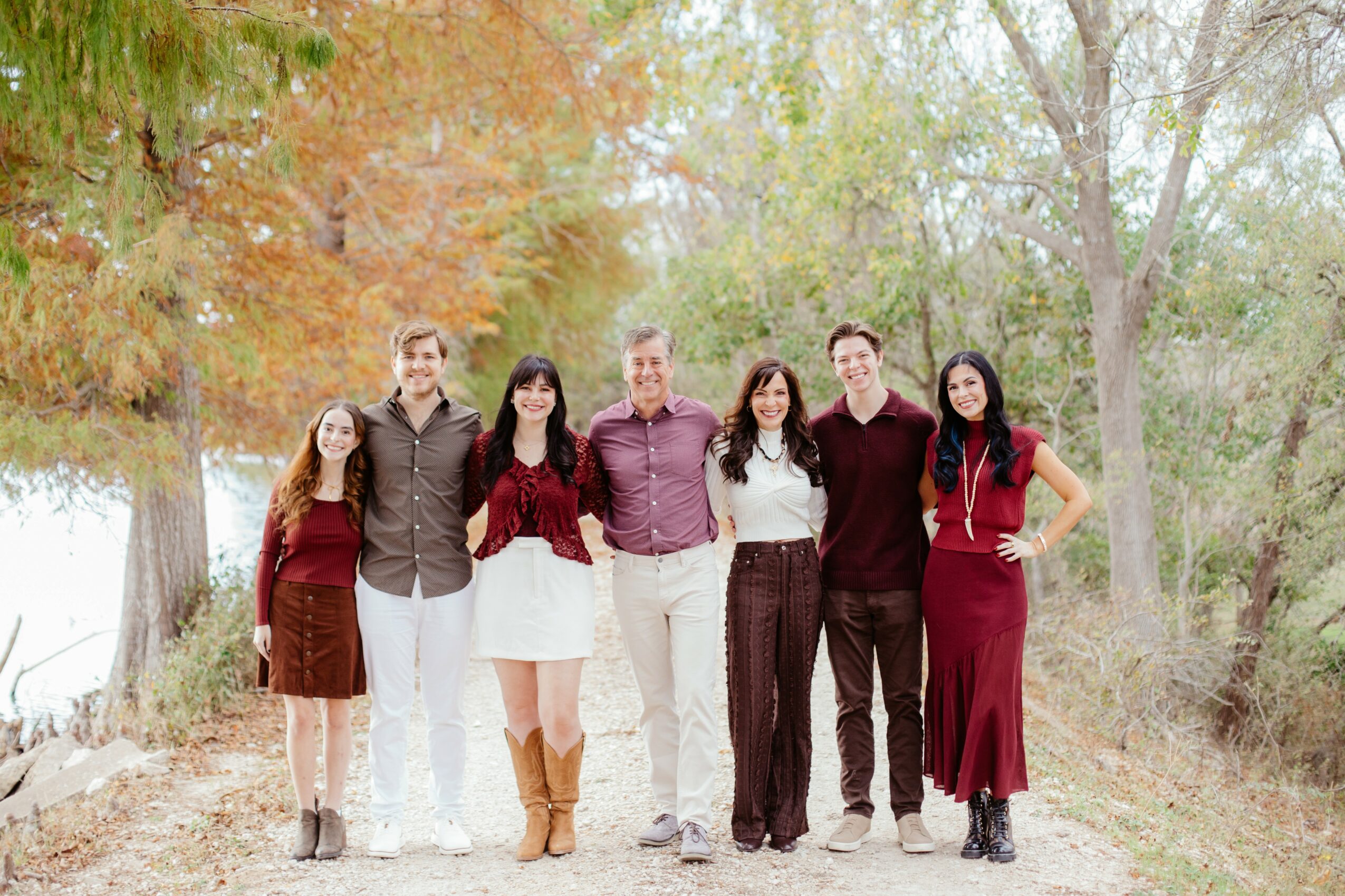 Group of seven people standing outdoors on a path surrounded by trees with autumn foliage, smiling at the camera.