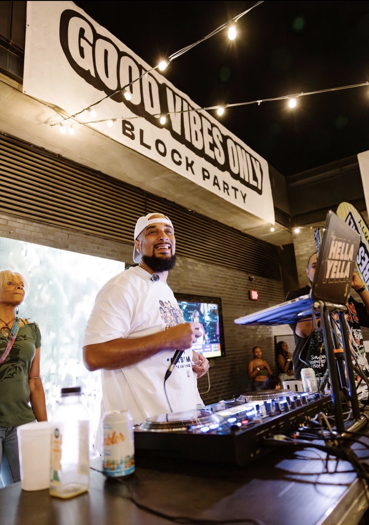 Man with a beard and white shirt behind DJ equipment at night, illuminated by string lights, with a sign reading 'Good Vibes Only Block Party'.