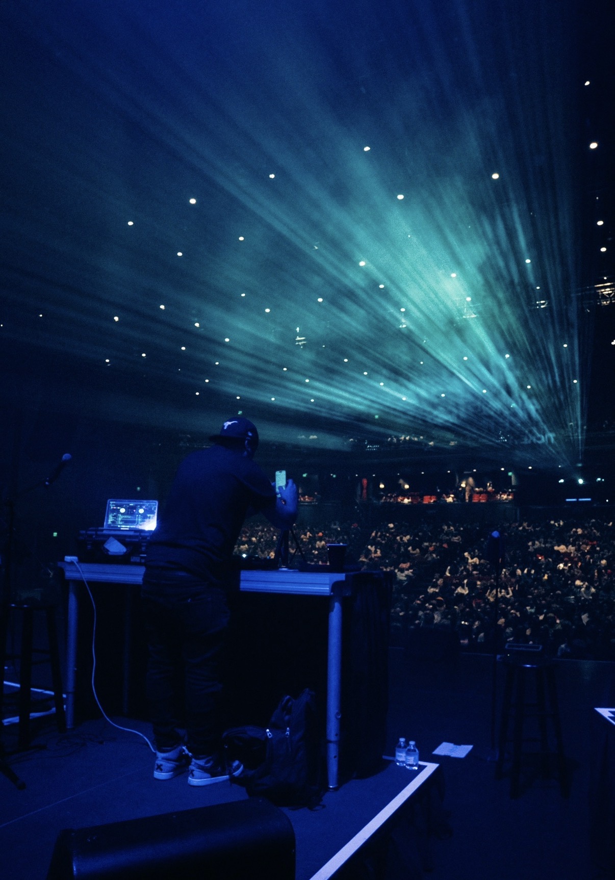 DJ performing on stage with bright blue and green light beams in a dark concert venue.