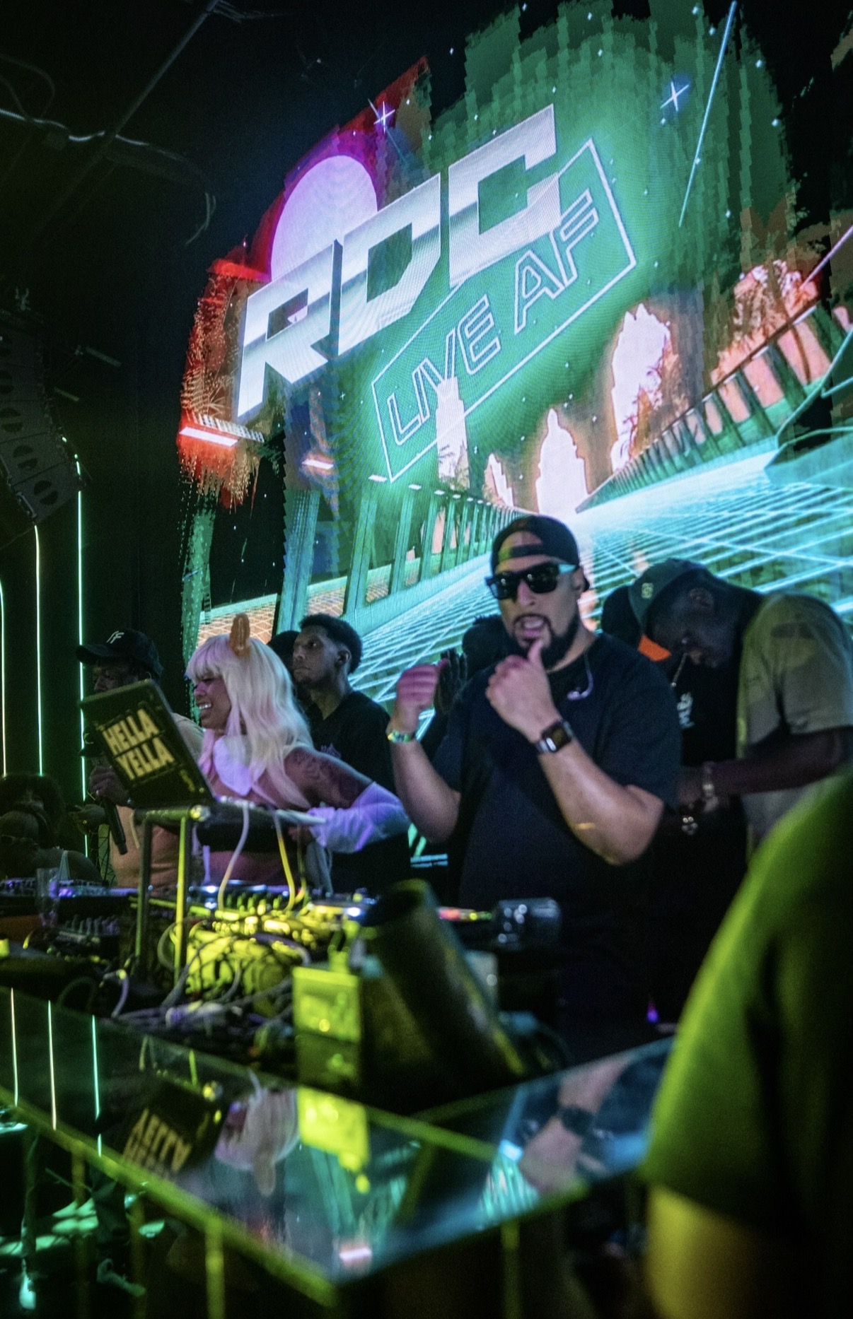 DJ wearing sunglasses and headphones performs at a nightclub with a colorful LED screen displaying 'RSC WEAR' behind him.